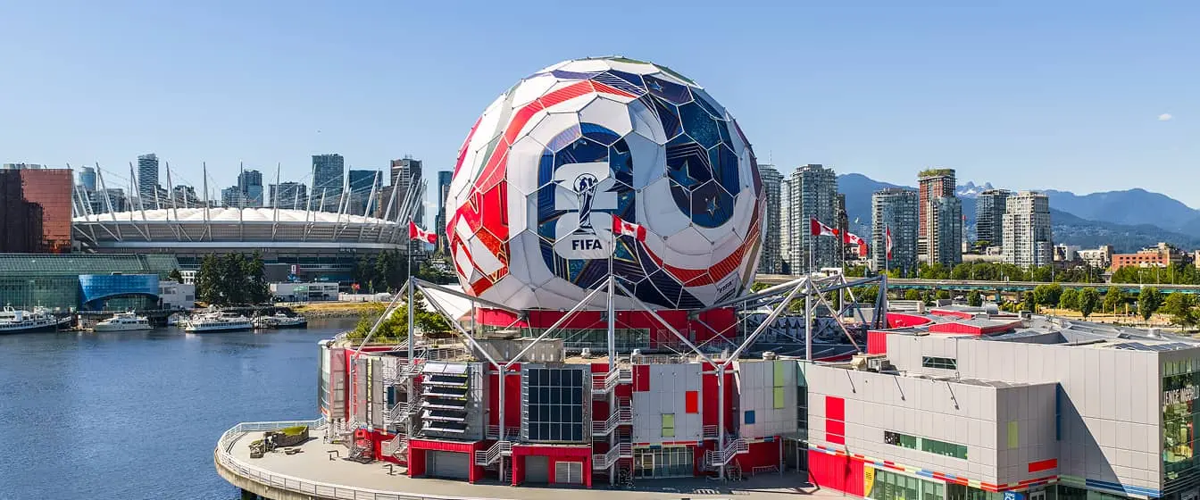 Large spherical building painted like a soccer ball with FIFA logo, surrounded by water and city skyline in the background.