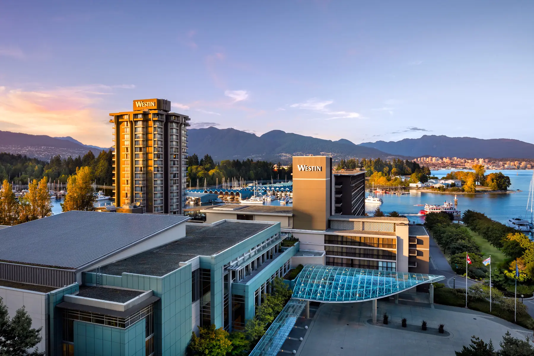 Westin hotel buildings near a marina with boats, trees with autumn colors, mountains, and a body of water at sunset.