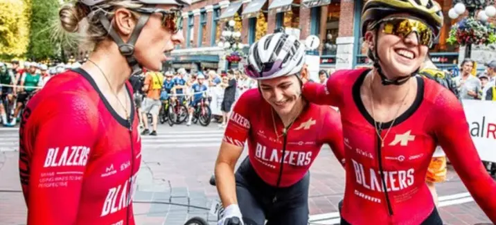 Three cyclists in red Blazers jerseys and helmets smiling and interacting on a city street with a crowd in the background.