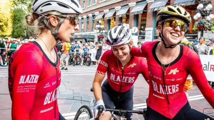 Three cyclists in red Blazers jerseys and helmets smiling and interacting on a city street with a crowd in the background.