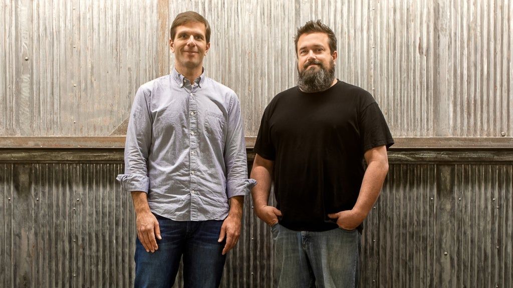 Two people standing in front of a corrugated metal wall, one wearing a light button-up shirt and jeans, the other in a black t-shirt and jeans.