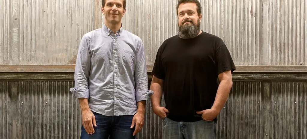 Two people standing in front of a corrugated metal wall, one wearing a light button-up shirt and jeans, the other in a black t-shirt and jeans.