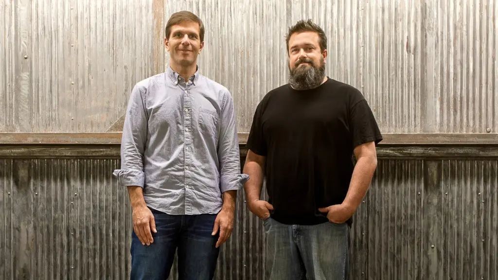 Two people standing in front of a corrugated metal wall, one wearing a light button-up shirt and jeans, the other in a black t-shirt and jeans.