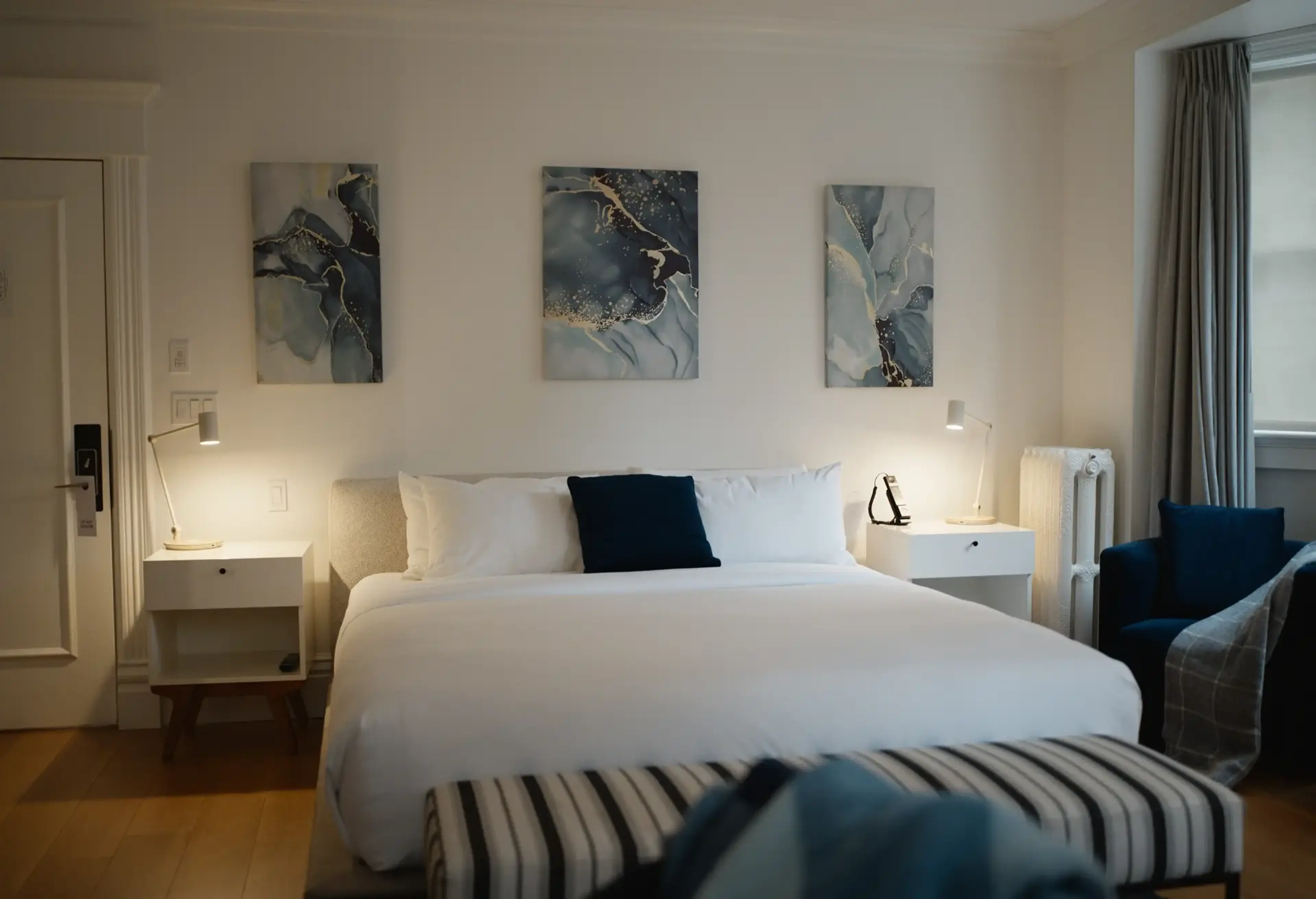 Modern bedroom with a large white bed, two white nightstands with lamps, three abstract paintings above the bed, and a blue armchair near a window with gray curtains.