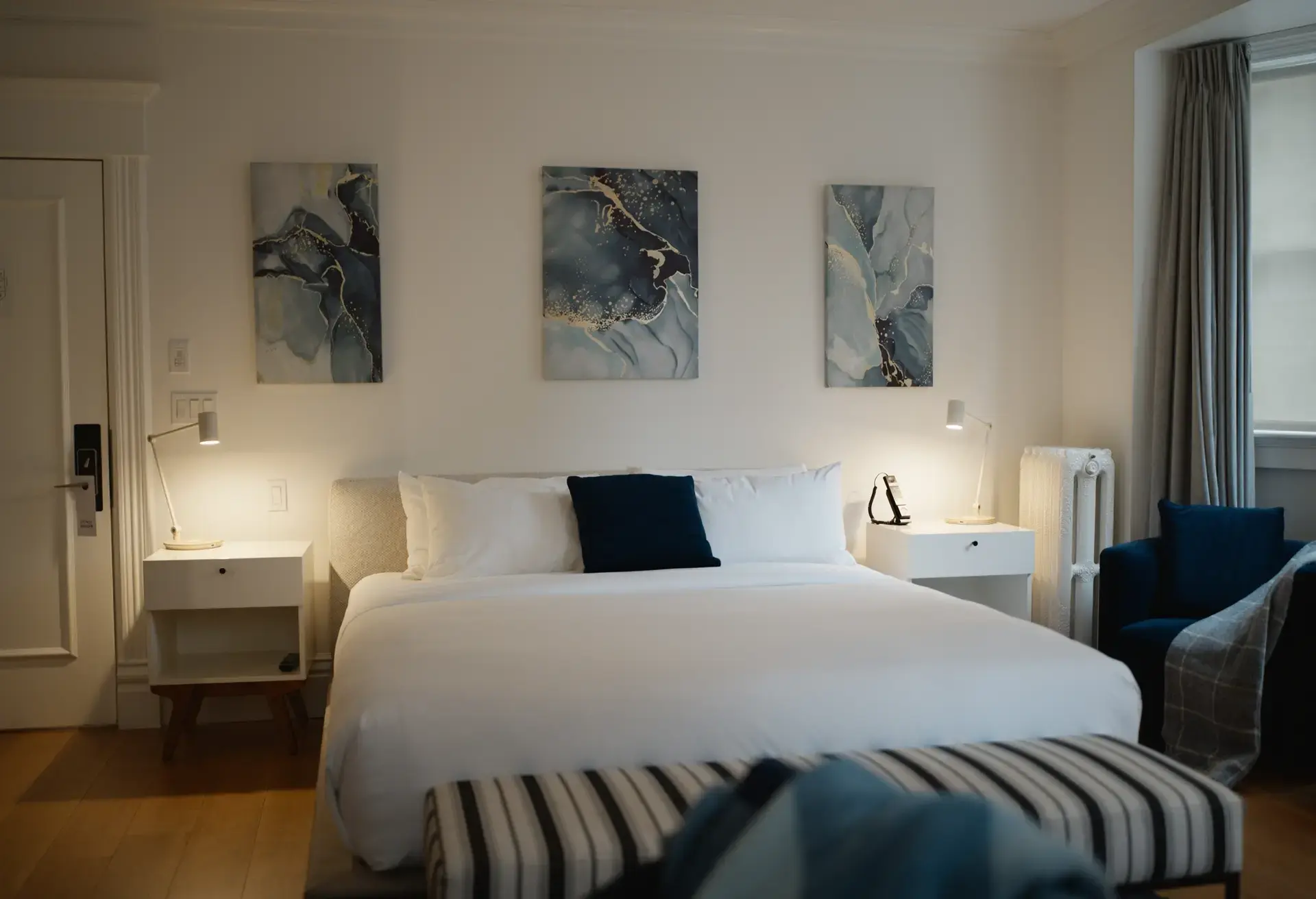 Modern bedroom with a large white bed, two white nightstands with lamps, three abstract paintings above the bed, and a blue armchair near a window with gray curtains.