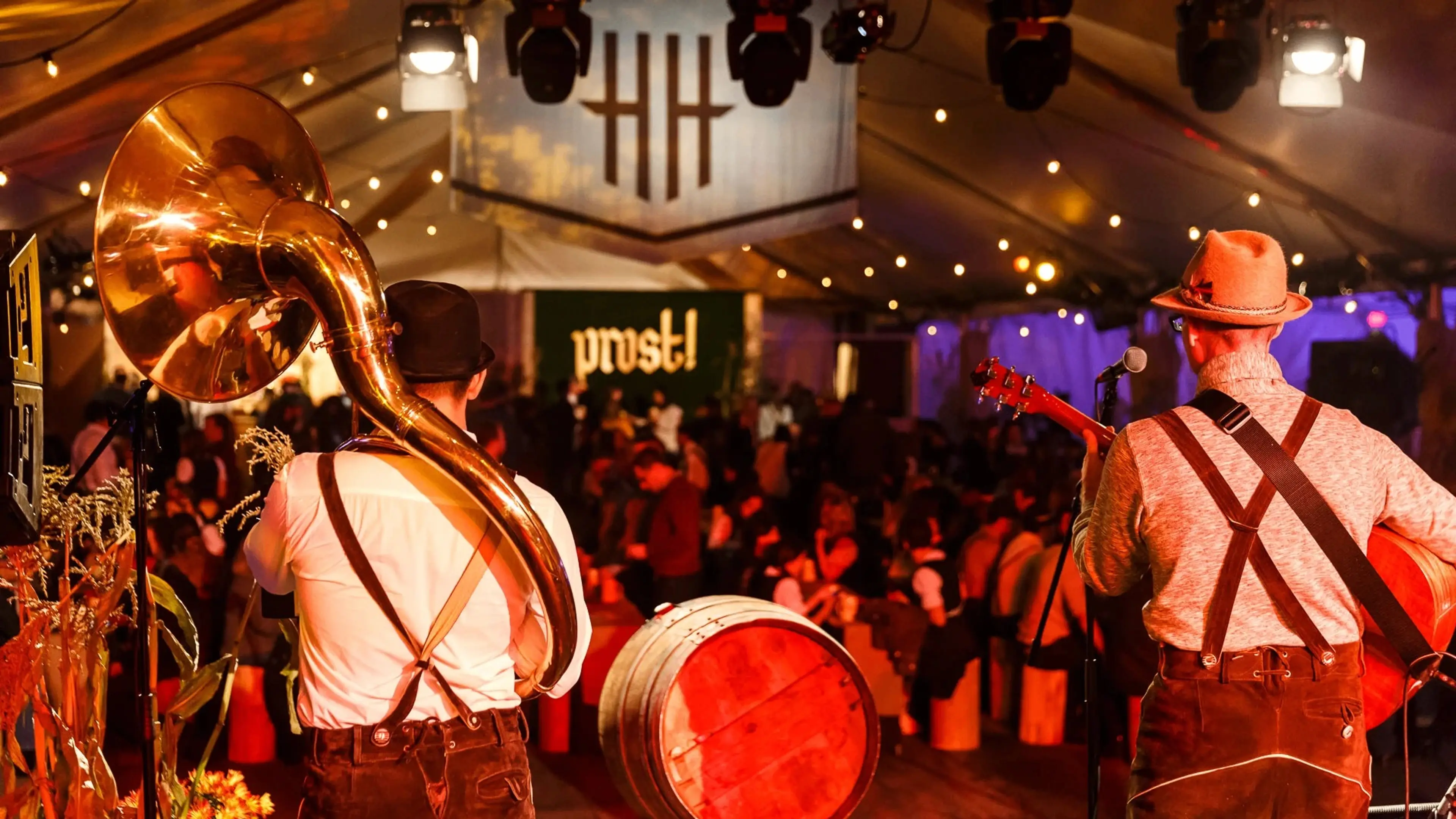 Two musicians in traditional attire playing sousaphone and guitar on stage at a festive indoor event with a crowd and 'prost!' sign in the background.