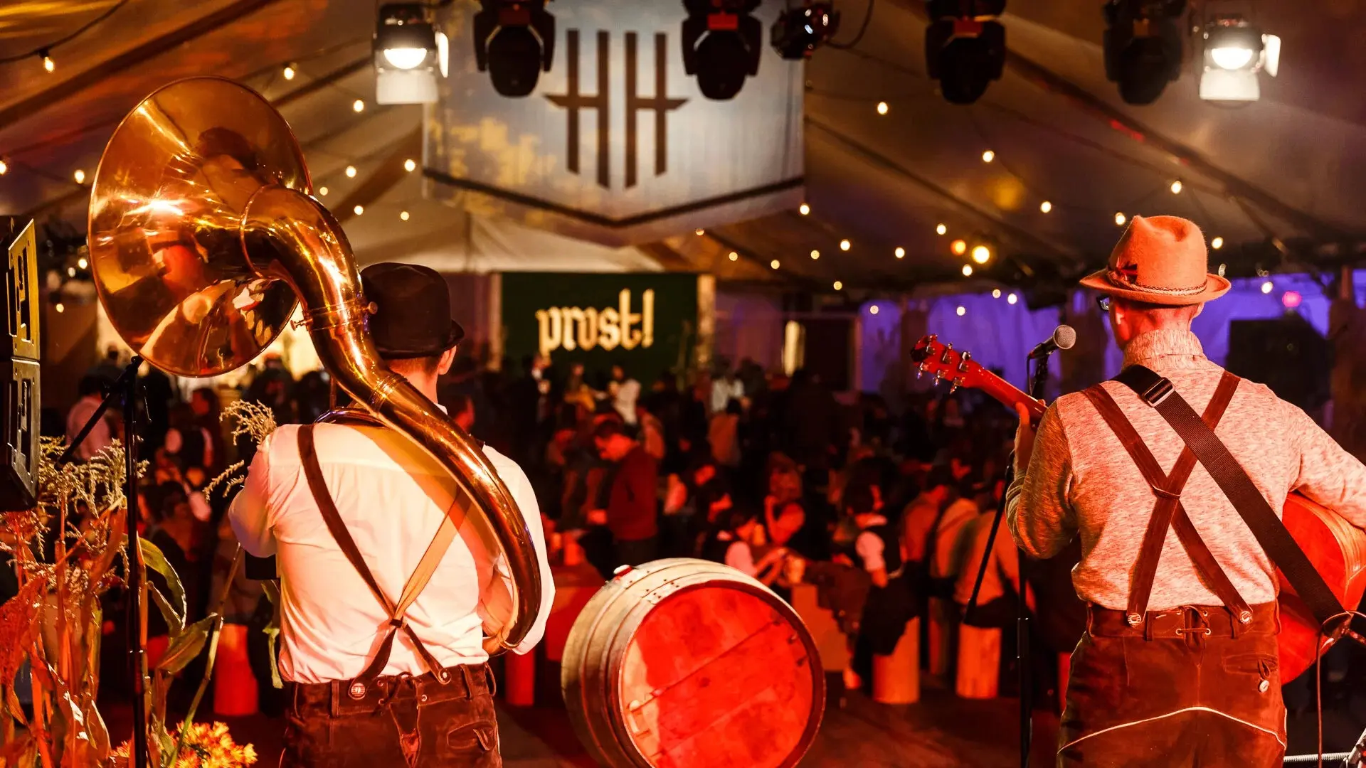 Two musicians in traditional attire playing sousaphone and guitar on stage at a festive indoor event with a crowd and 'prost!' sign in the background.