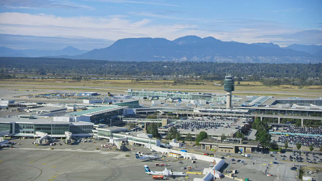 Aerial view of a large airport with multiple terminals, parked airplanes, control tower, and surrounding parking lots with mountains in the background.