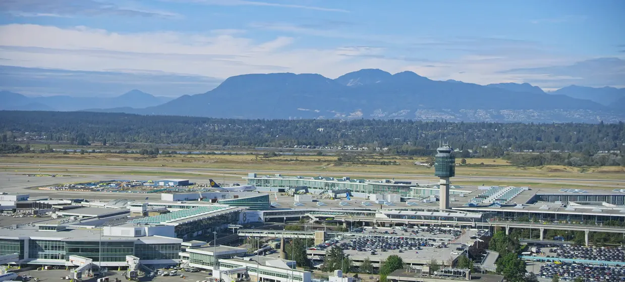 Aerial view of a large airport with multiple terminals, parked airplanes, control tower, and surrounding parking lots with mountains in the background.