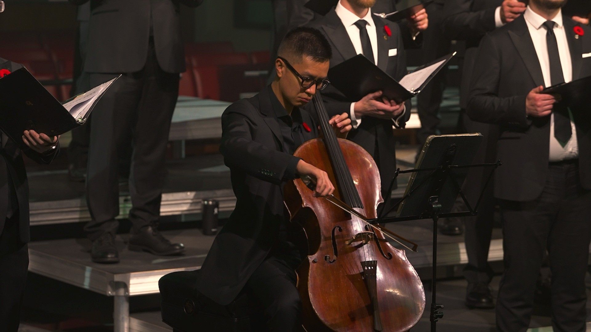 A person in black suit playing cello on stage with a group of people in black suits holding music folders.