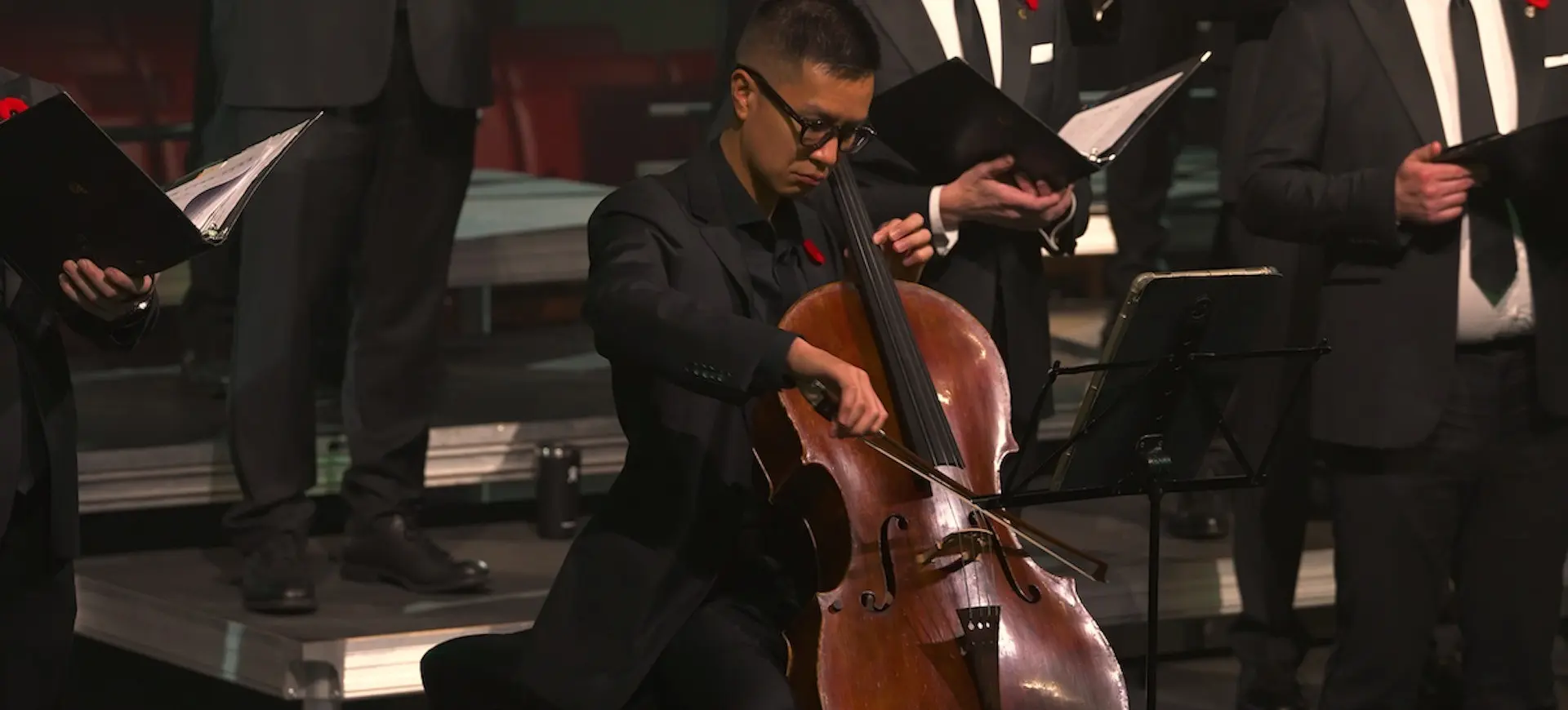 A person in black suit playing cello on stage with a group of people in black suits holding music folders.