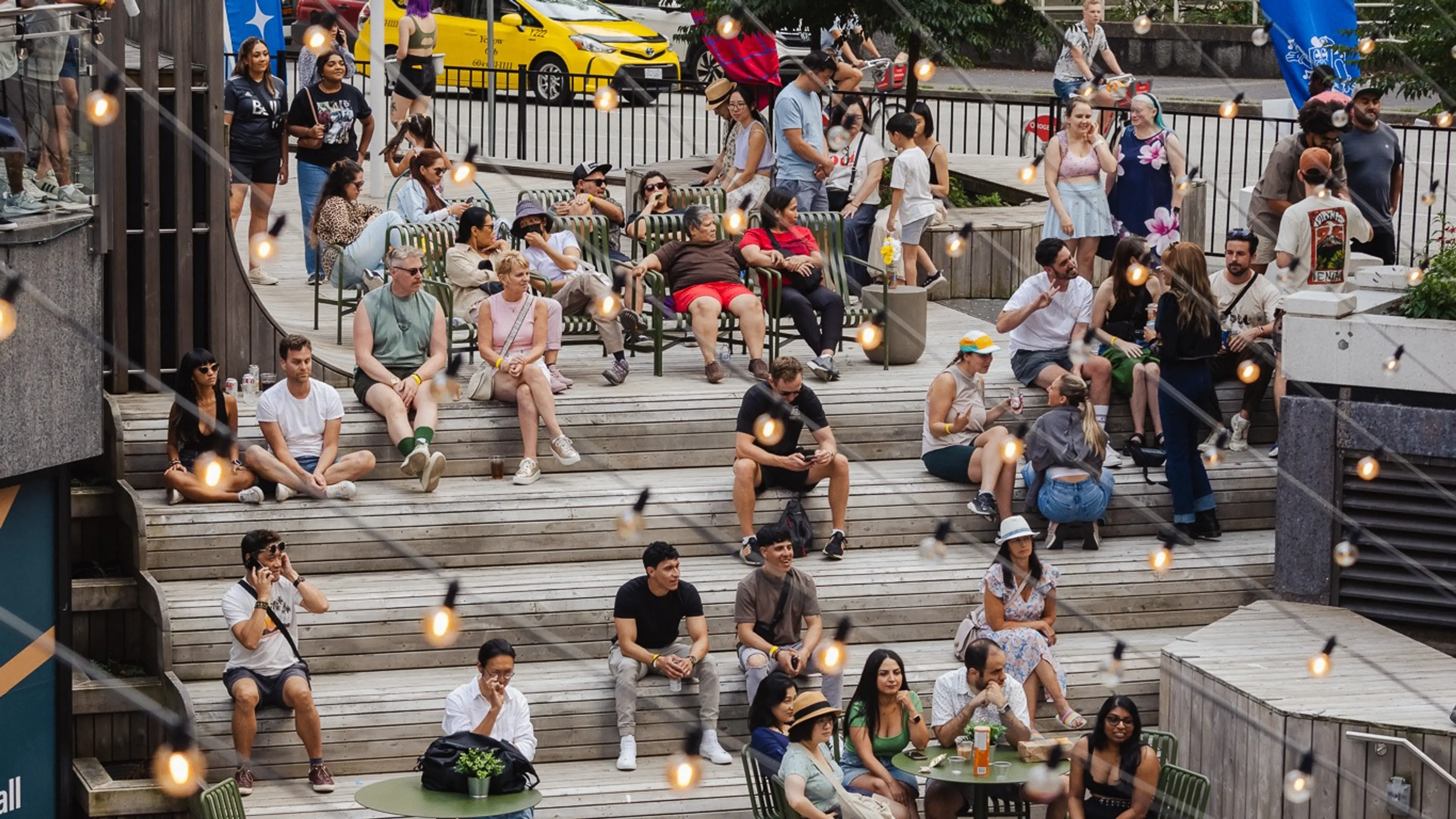 Crowd of people sitting and standing on outdoor wooden steps and benches with string lights hanging above during daytime.