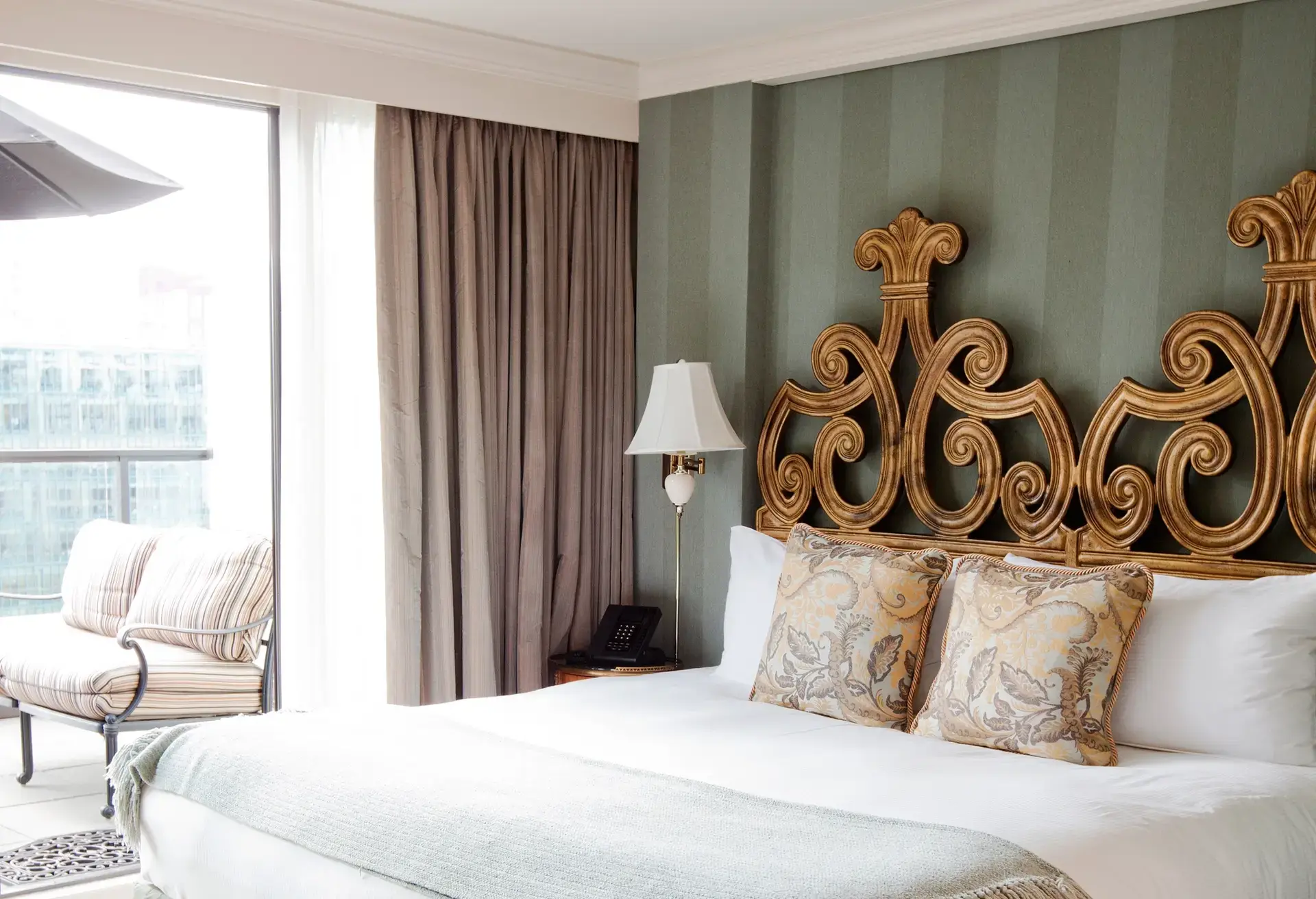 Bed with ornate gold headboard, two patterned pillows, white bedding, bedside lamp, and balcony with cushioned chair.