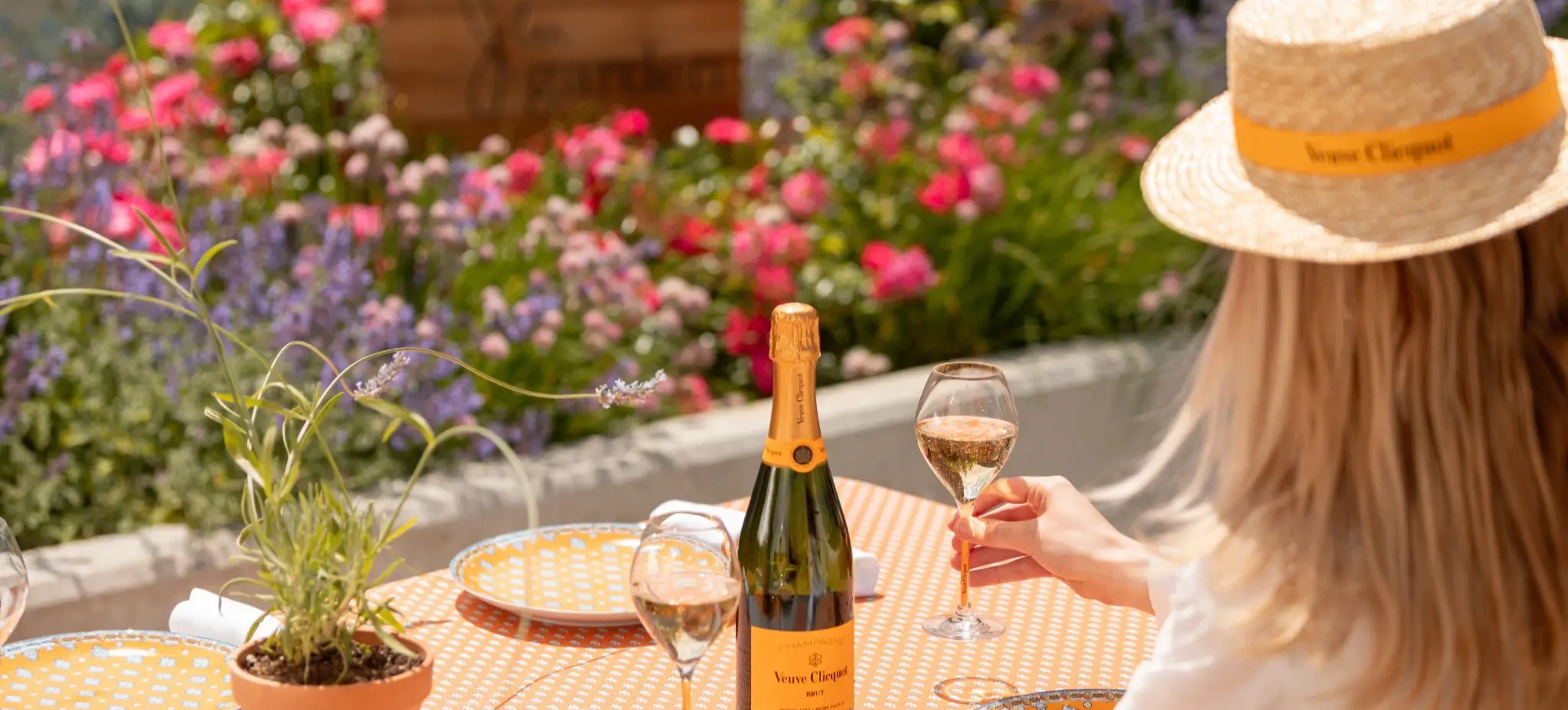 Person wearing a straw hat holding a glass of champagne at a table with a champagne bottle, two glasses, and a potted plant, with colorful flowers in the background.