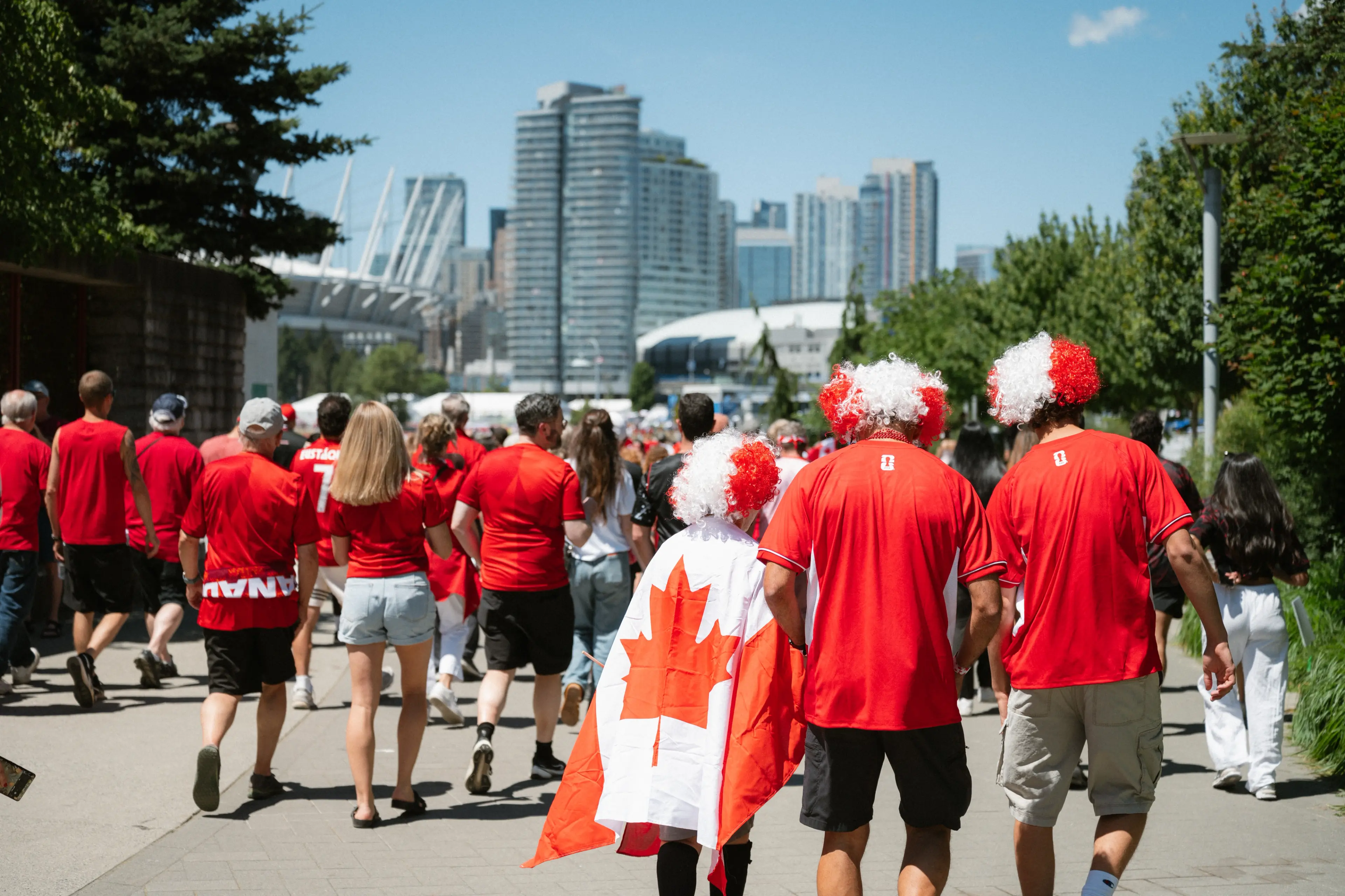 Crowd of people walking outdoors wearing red shirts, with three individuals wearing red and white wigs and one draped in a Canadian flag.