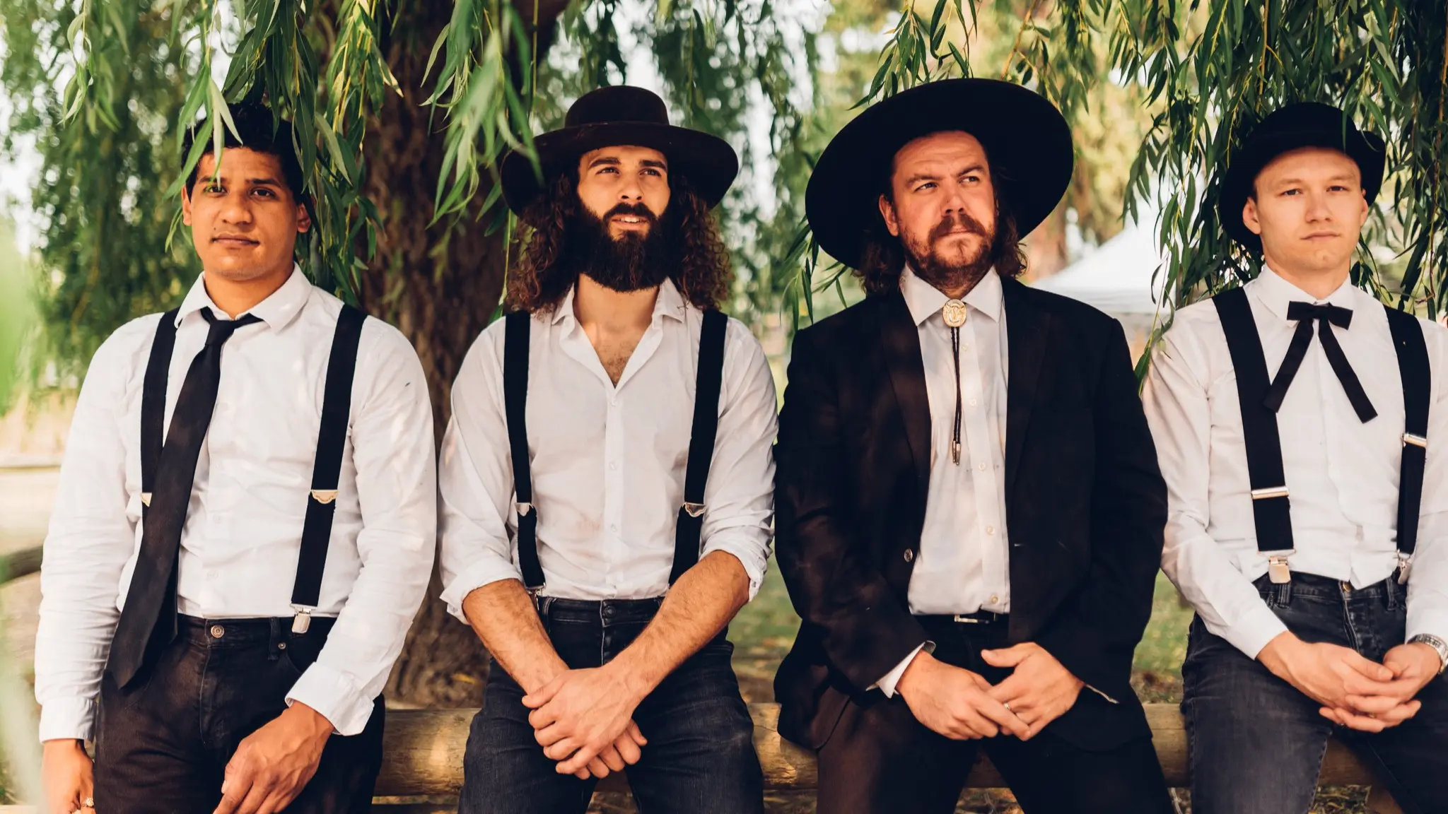 Four people wearing white shirts, black suspenders, and black hats standing outdoors under tree branches.