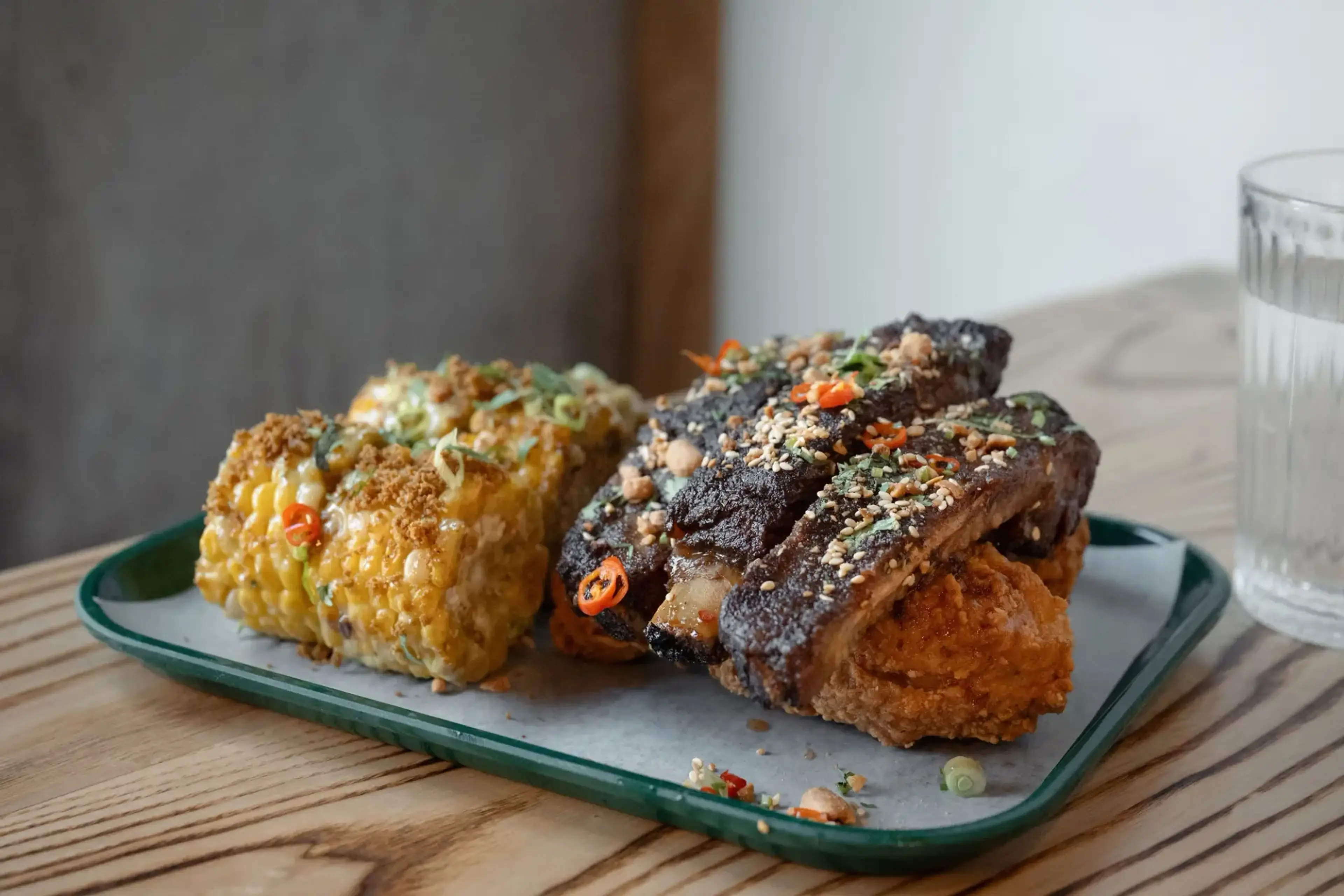 Plate with grilled corn on the cob topped with herbs and spices, and fried chicken ribs garnished with sesame seeds and chili slices.