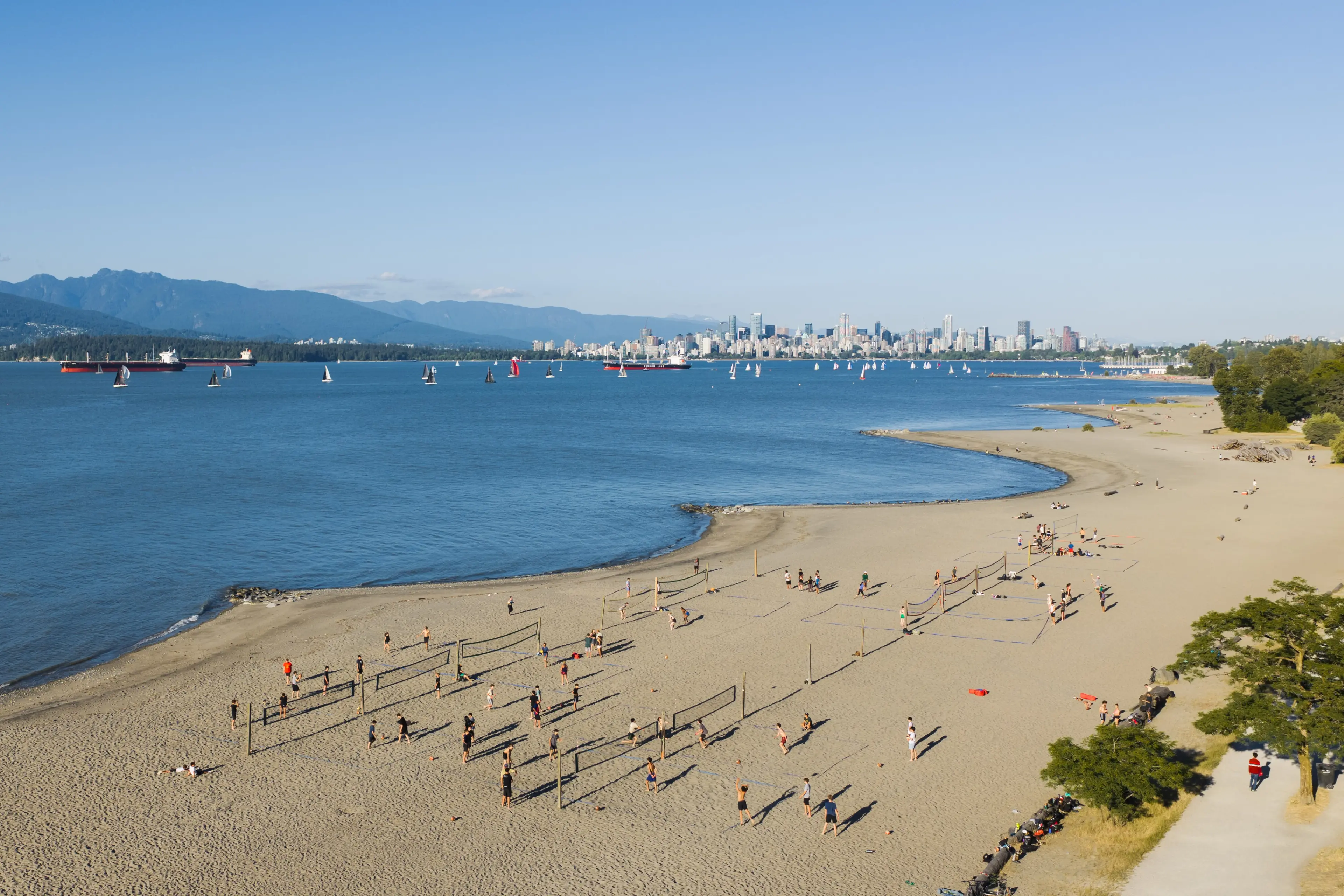 Spanish Banks Beach aerial view with multiple volleyball courts and people playing, sailboats on water, city skyline and mountains in the background.
