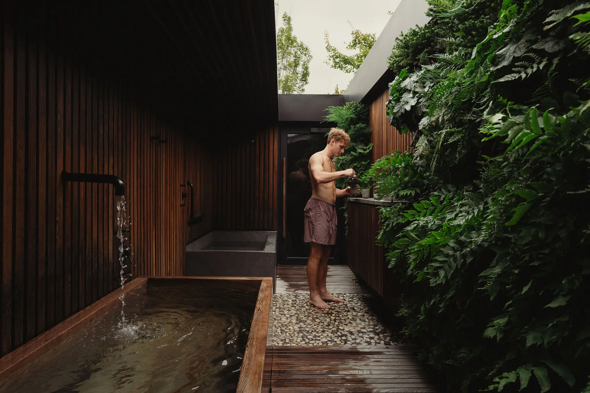 A person pouring water into a cup standing in the middle of an outdoor wellness retreat. Water is filling a wooden bathtub.
