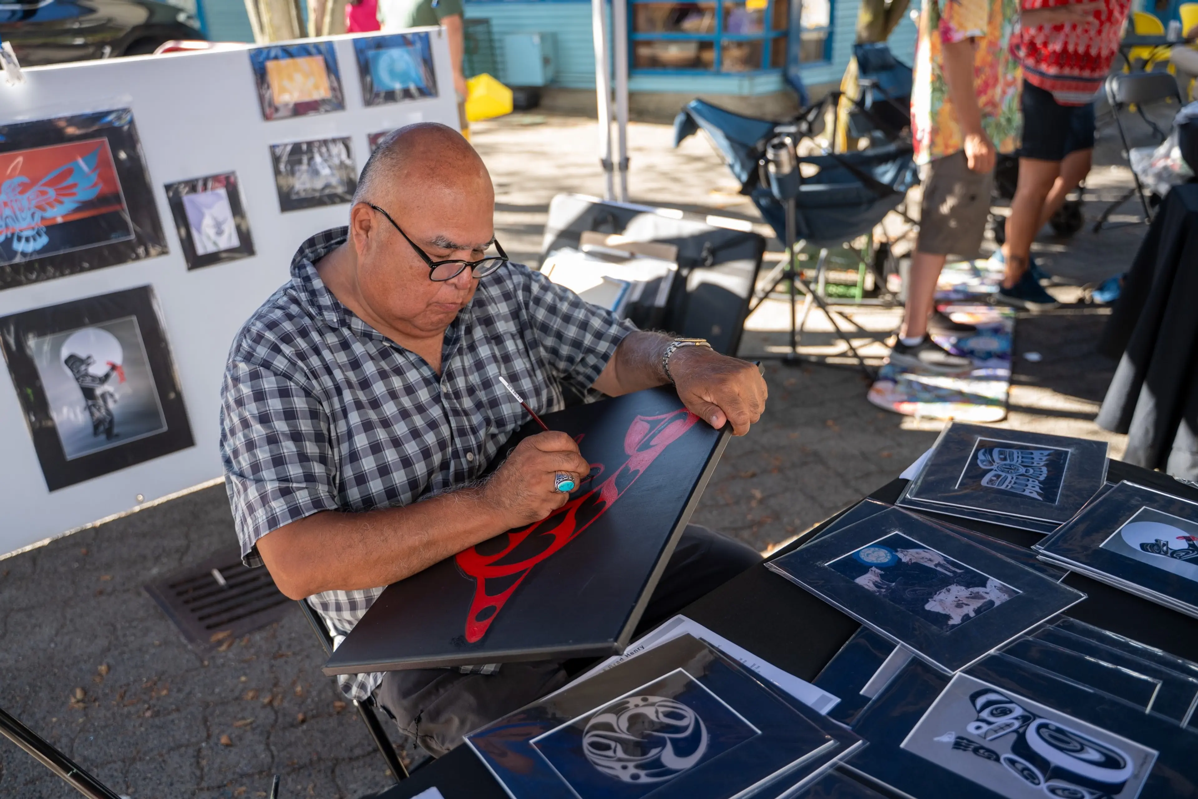 A person wearing glasses and a checkered shirt painting a red design on a black canvas at an outdoor art market.