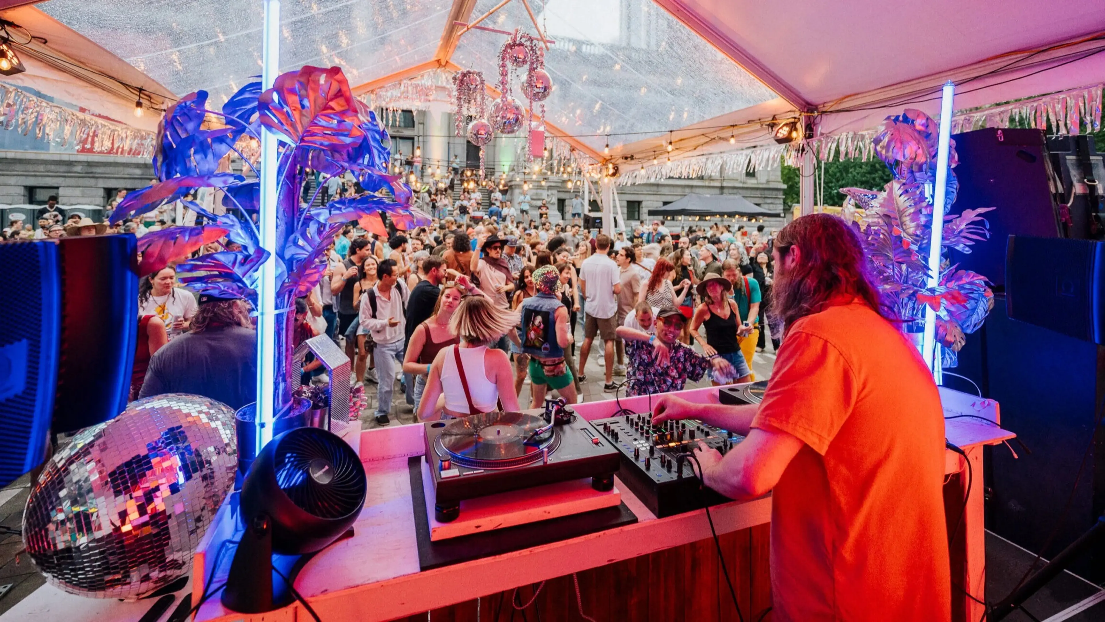 DJ in orange shirt playing music on turntables under a tent with colorful lights, crowd dancing outside.