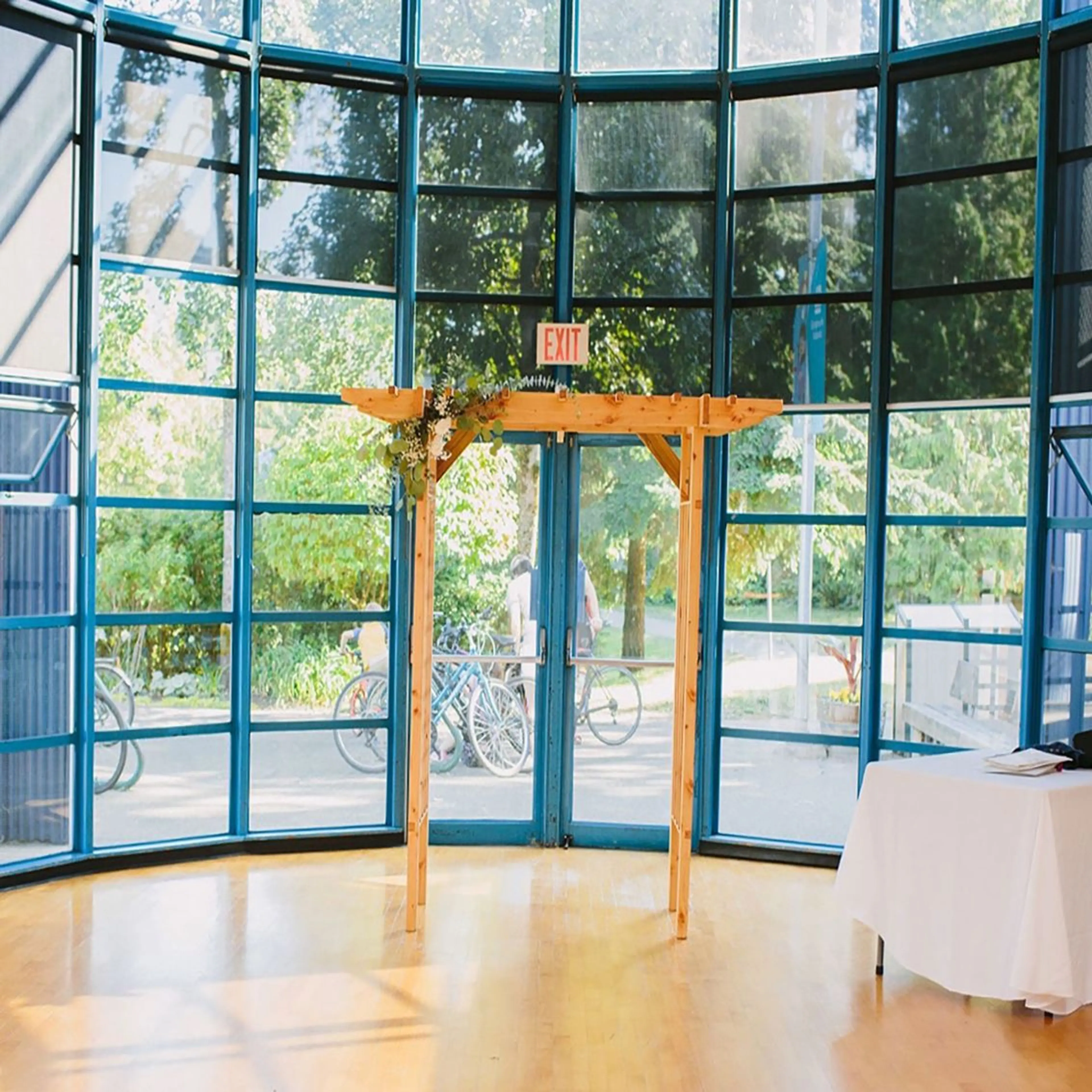 Wooden arch decorated with flowers inside a room with large blue-framed windows and a table with a white cloth.