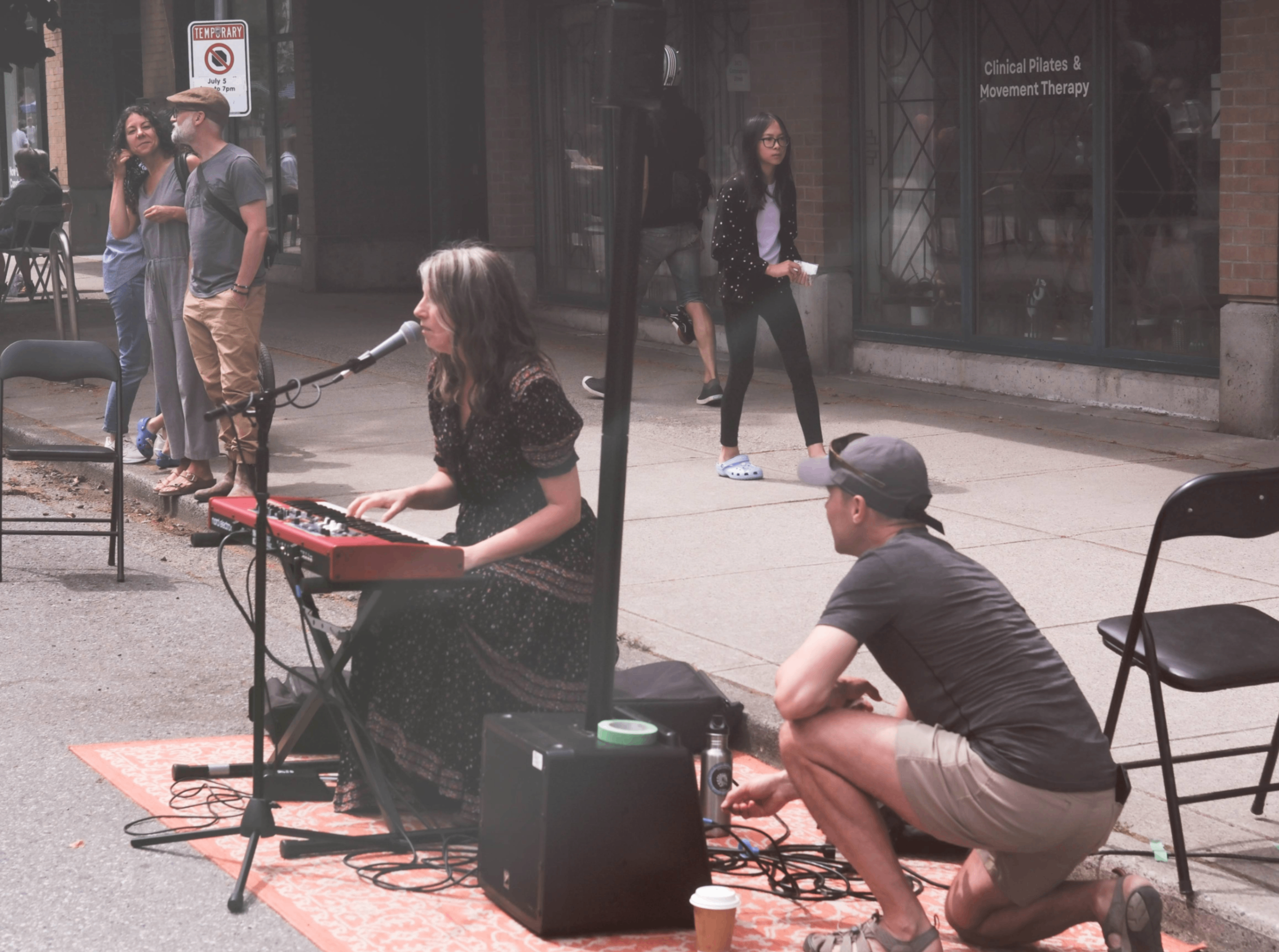 Person playing keyboard and singing into microphone on street with another person crouching nearby adjusting equipment, and three people standing in background.
