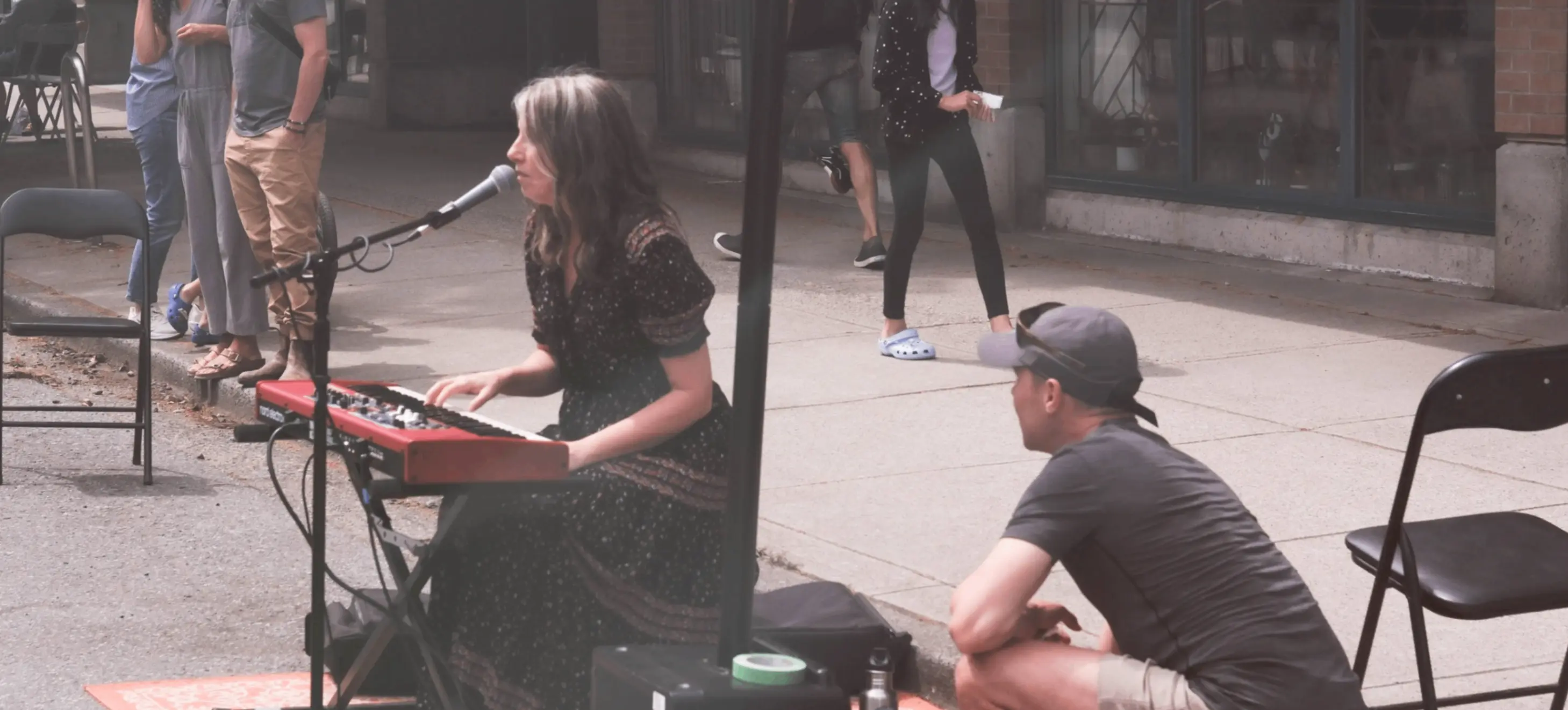 Person playing keyboard and singing into microphone on street with another person crouching nearby adjusting equipment, and three people standing in background.