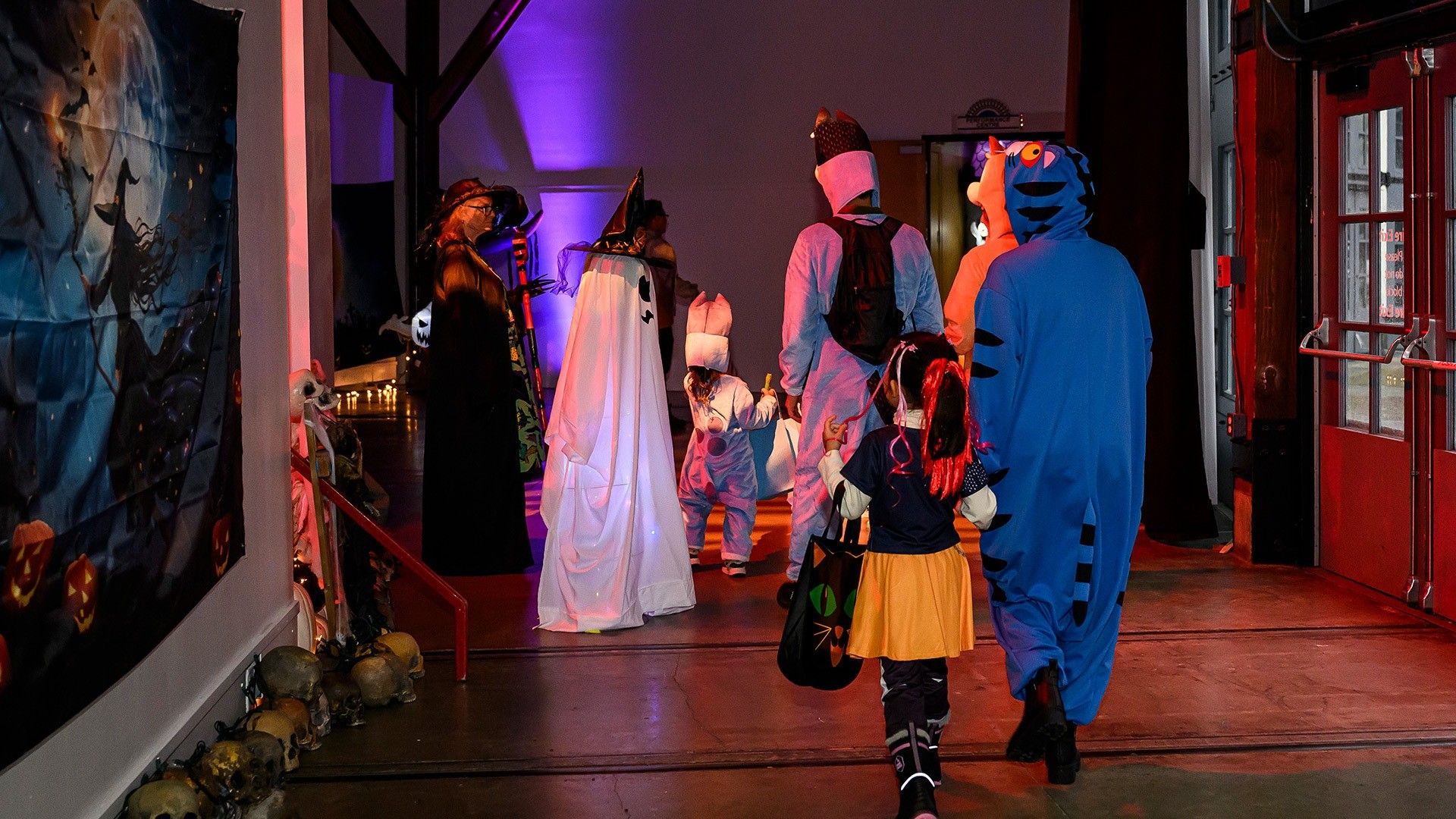 Group of people and children in Halloween costumes walking indoors toward a purple-lit area, including a ghost, tiger, and witch.