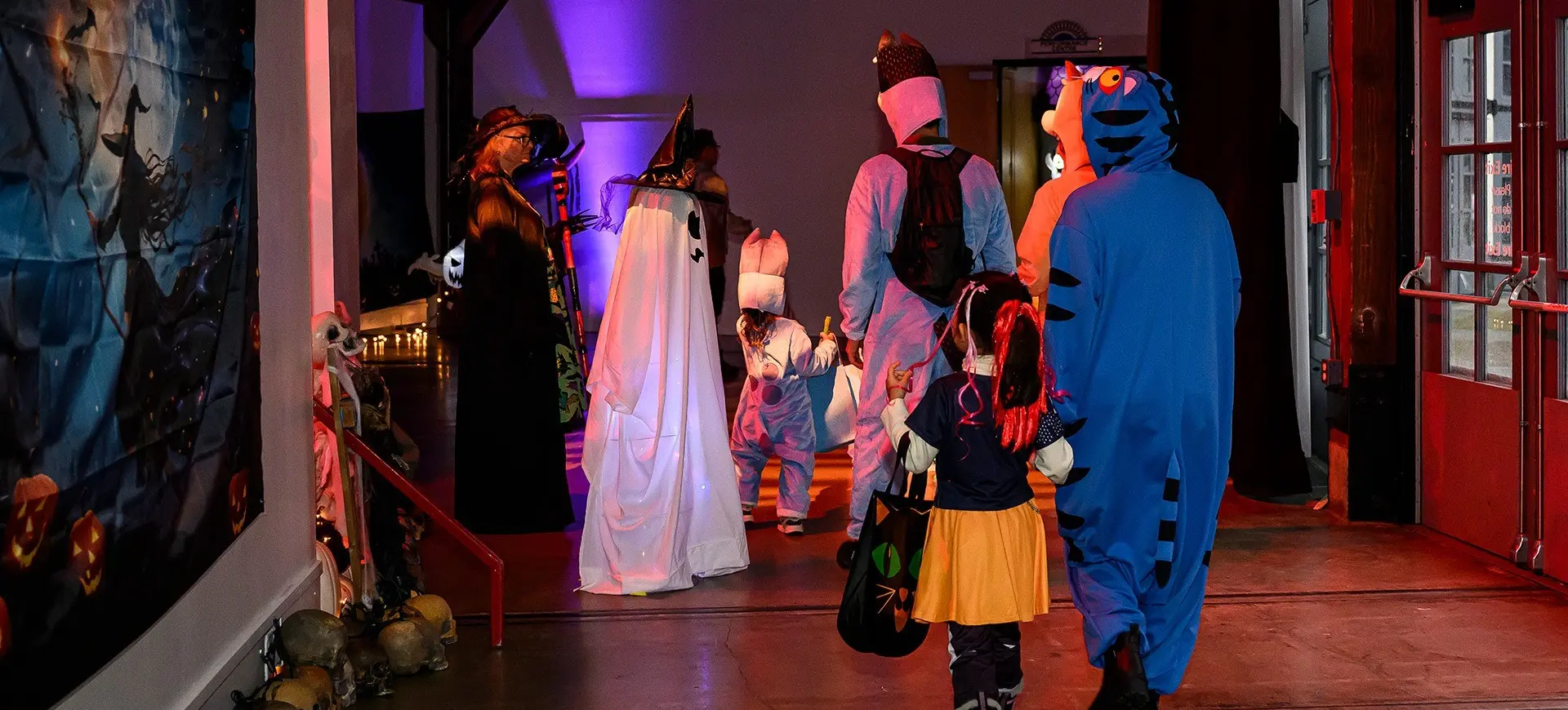 Group of people and children in Halloween costumes walking indoors toward a purple-lit area, including a ghost, tiger, and witch.