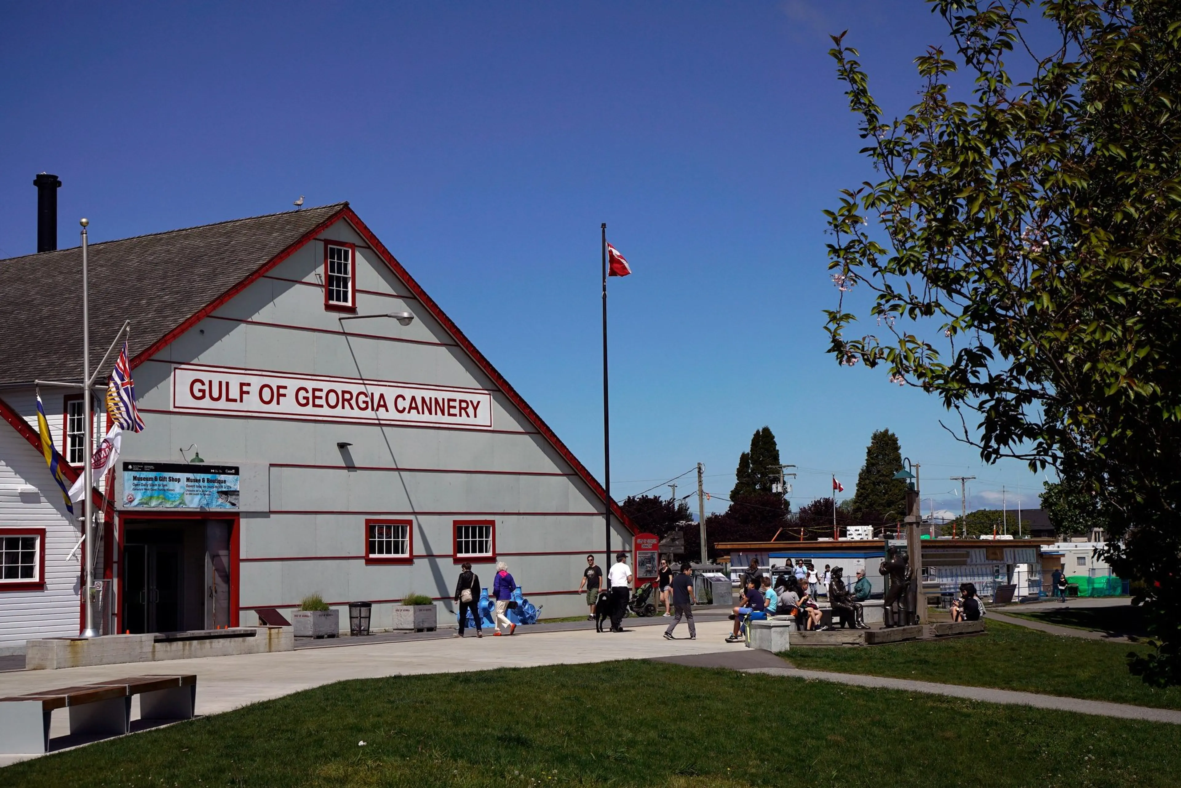 People walking and sitting near the Gulf of Georgia Cannery building under a clear blue sky with flags flying.