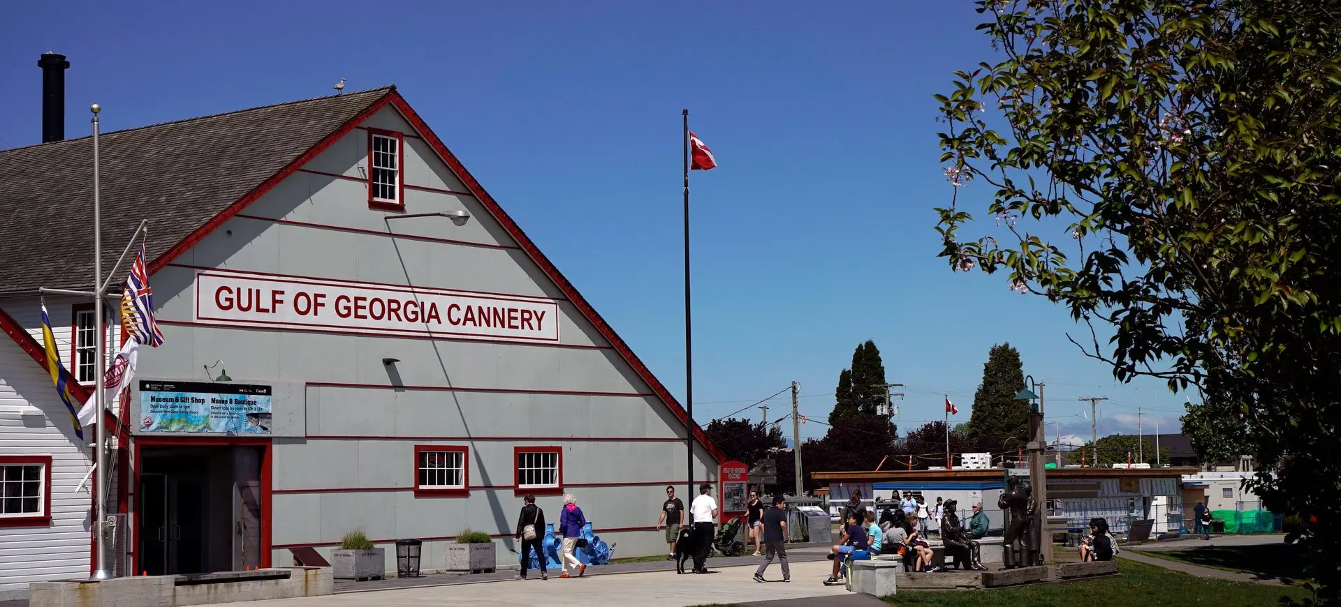 People walking and sitting near the Gulf of Georgia Cannery building under a clear blue sky with flags flying.