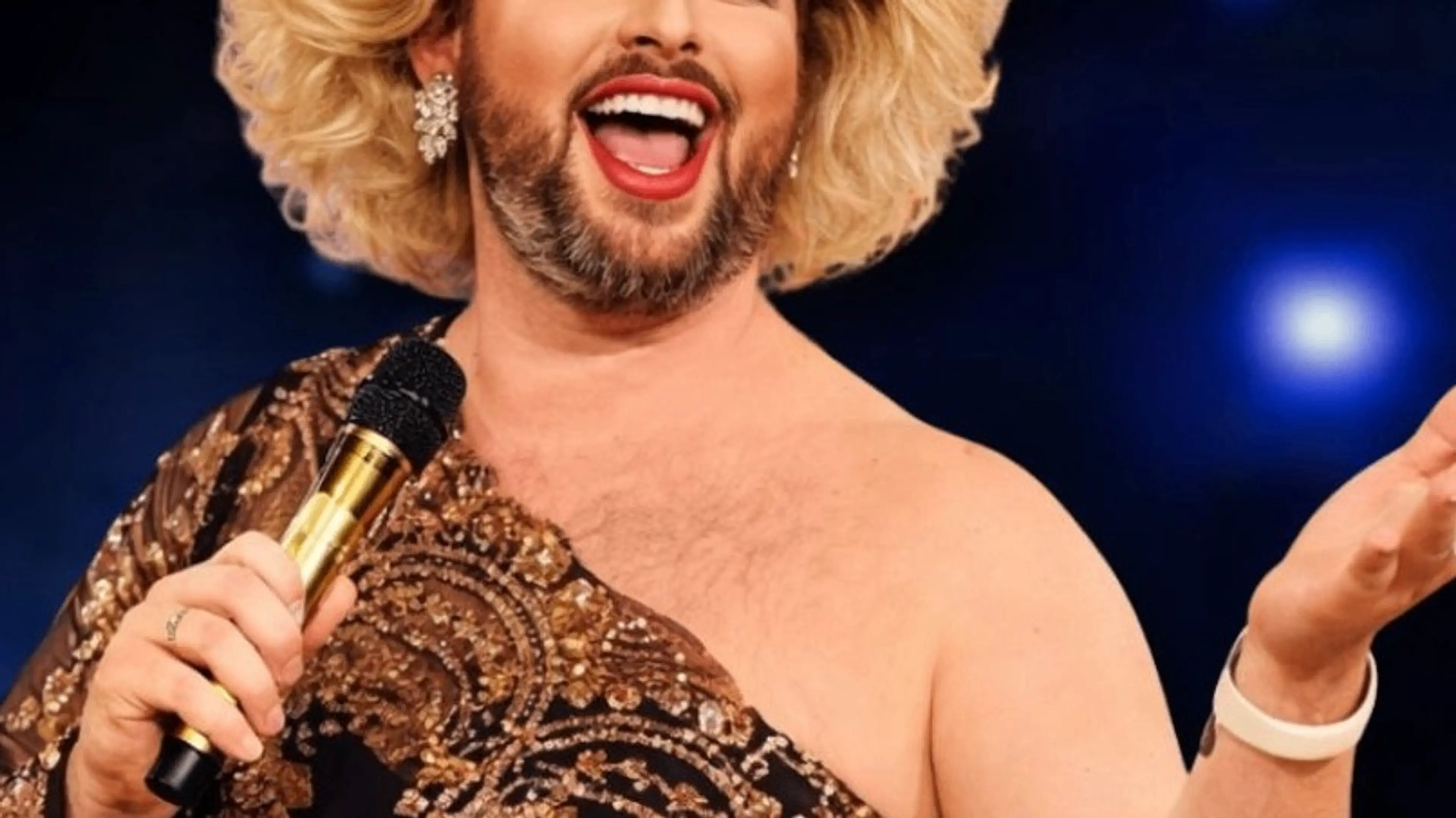 Person with blonde curly hair, beard, and red lipstick holding a microphone and smiling, wearing a sequined one-shoulder dress.