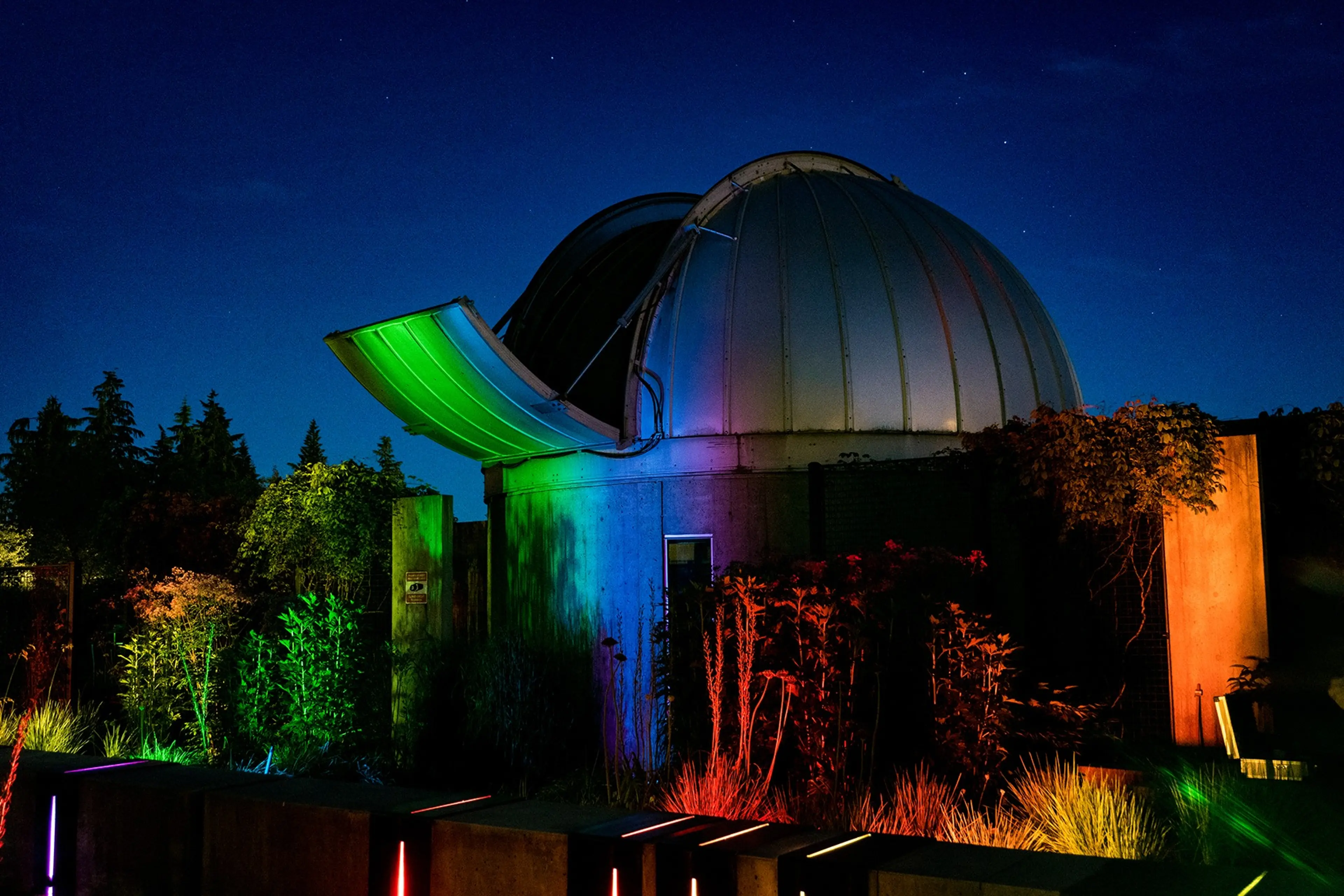The exterior of the SFU Trottier Observatory in Vancouver at night with the telescope door open and the building lit up with soft colourful lights