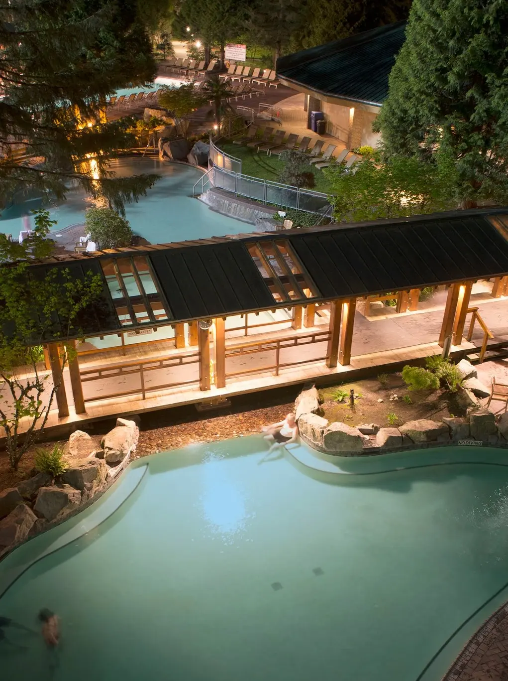 Night view of a lit outdoor pool area with people swimming and a covered walkway surrounded by trees and lounge chairs.