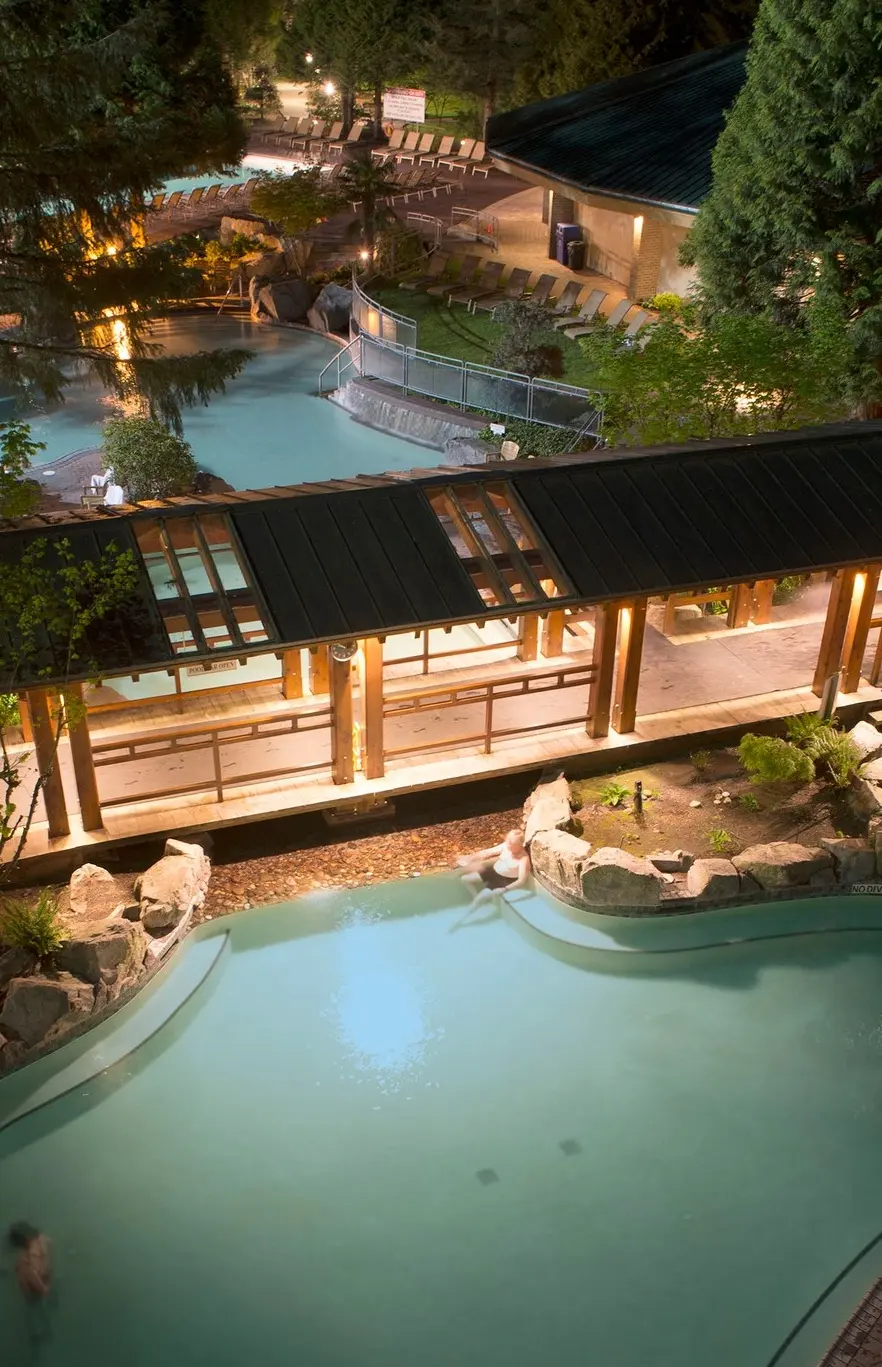 Night view of a lit outdoor pool area with people swimming and a covered walkway surrounded by trees and lounge chairs.