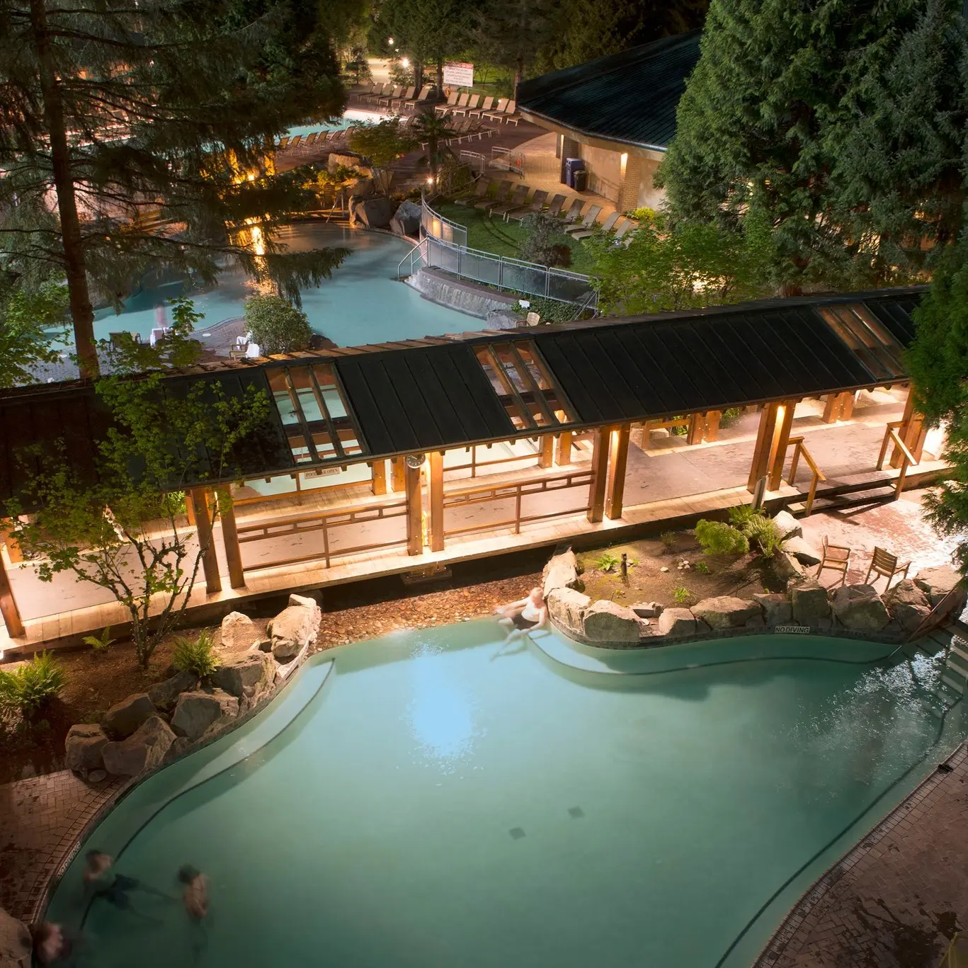 Night view of a lit outdoor pool area with people swimming and a covered walkway surrounded by trees and lounge chairs.