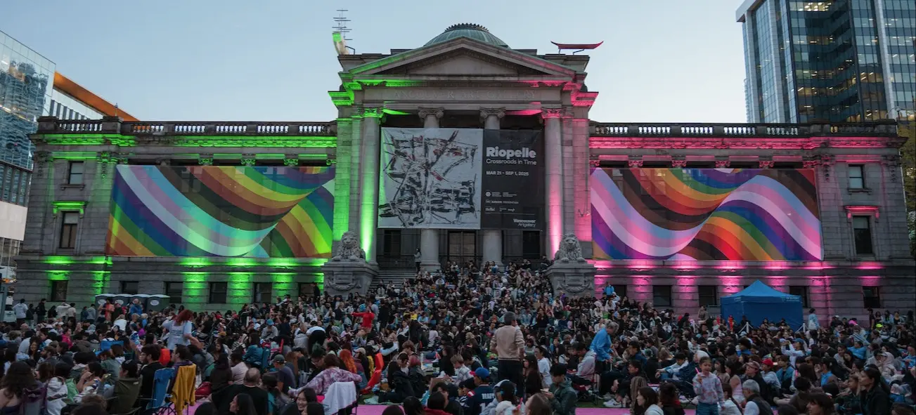 Large crowd gathered in front of a historic building with rainbow-colored light projections and banners for Riopelle exhibition.