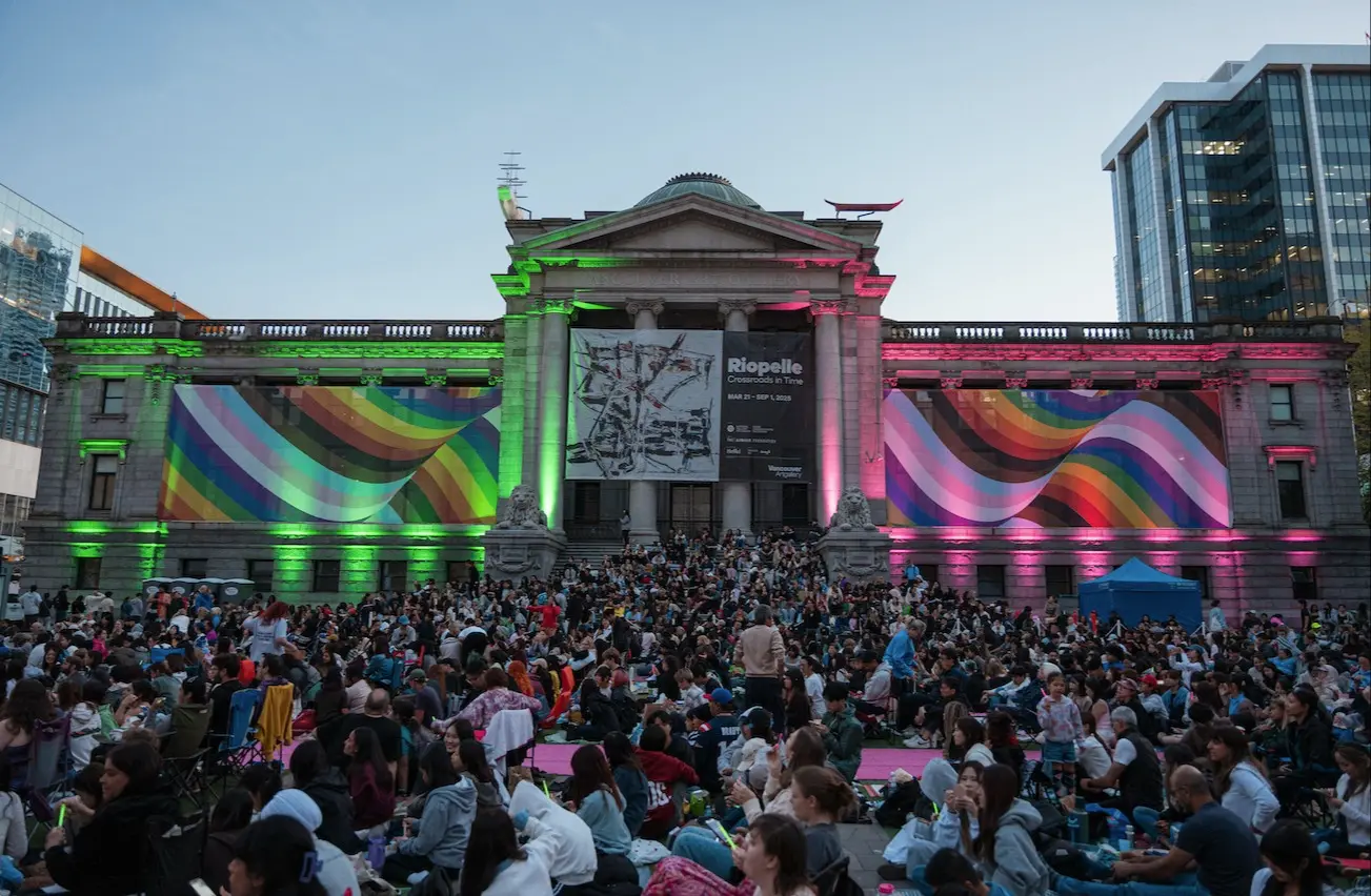 Large crowd gathered in front of a historic building with rainbow-colored light projections and banners for Riopelle exhibition.