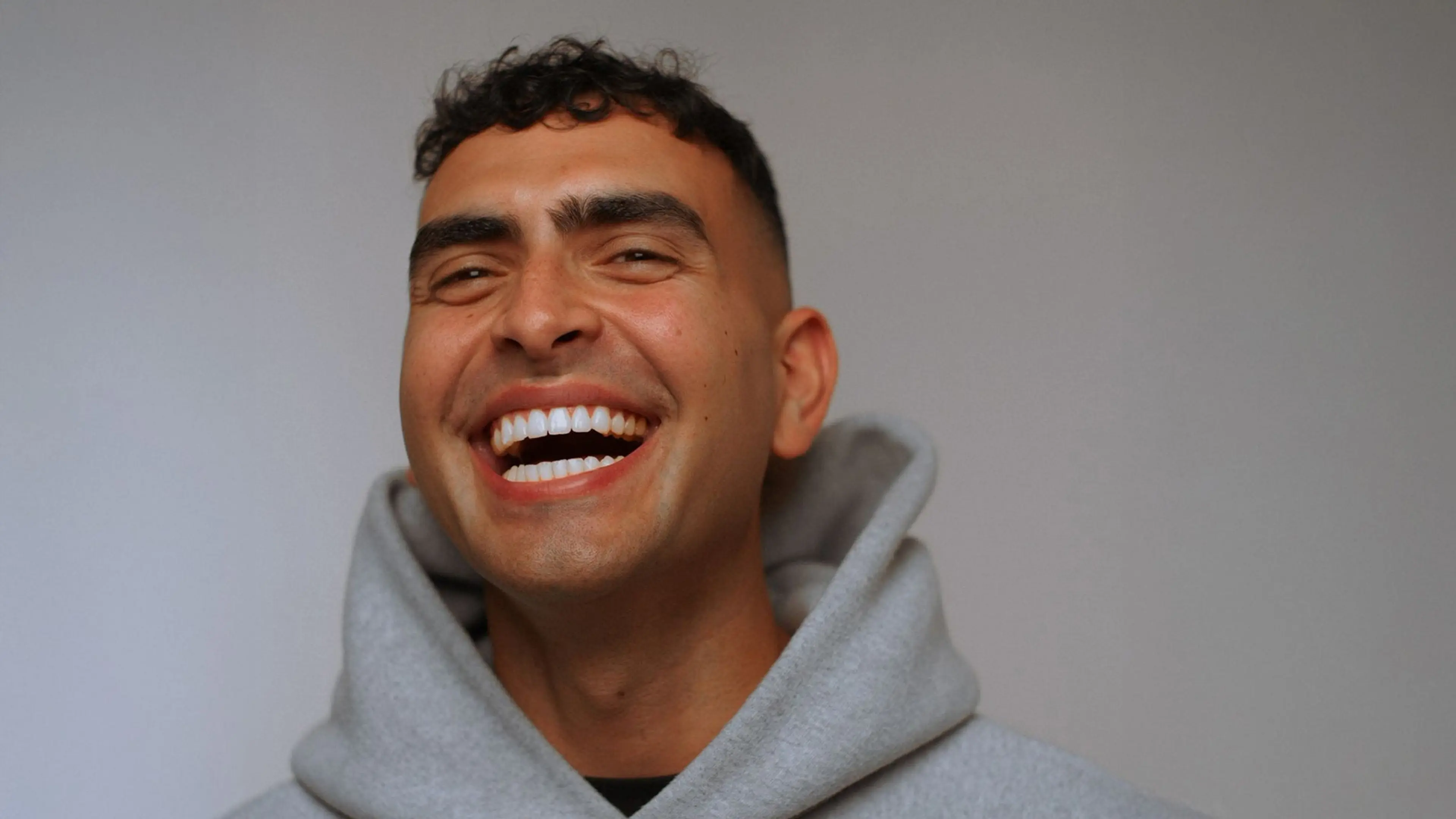 Person with short curly hair wearing a gray hoodie, smiling broadly against a plain background.