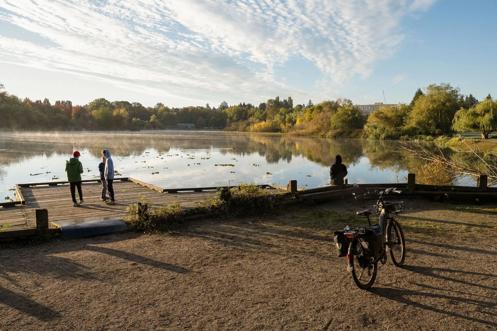 Two people standing on a wooden dock and one person sitting by a calm lake with trees and a bicycle nearby under a partly cloudy sky.