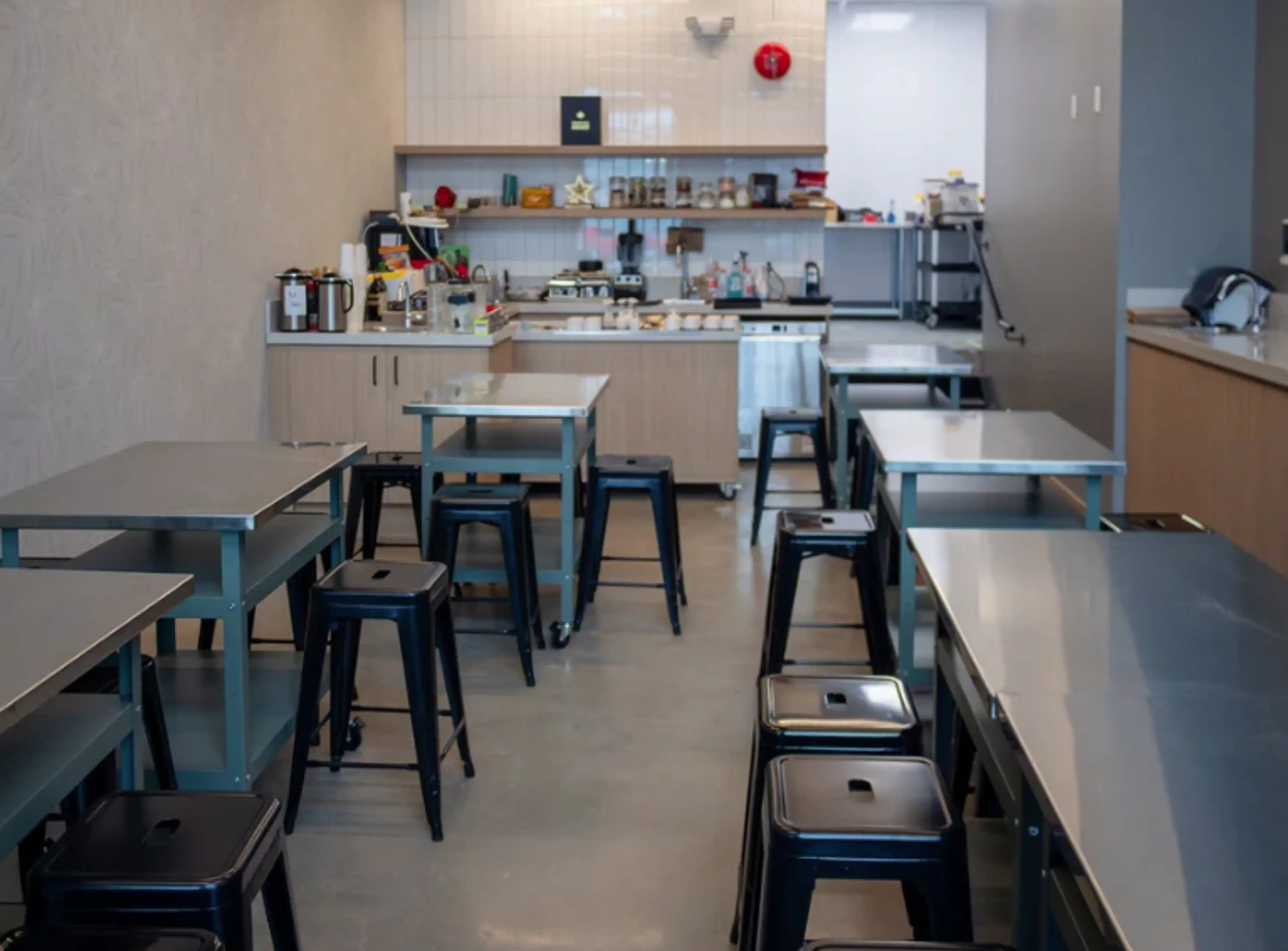 Empty kitchen with metal tables and black stools, shelves with jars and kitchen appliances in the background.