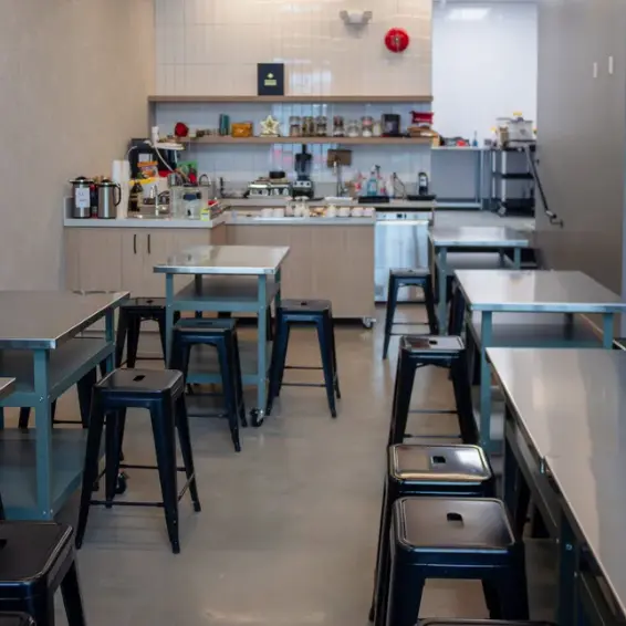 Empty kitchen with metal tables and black stools, shelves with jars and kitchen appliances in the background.