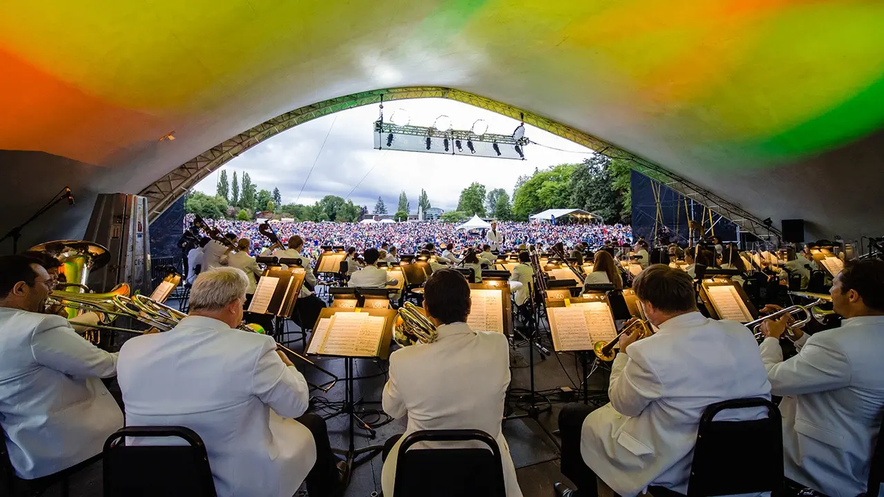 Orchestra performing under a large tent with colorful lighting, facing a large outdoor audience in a park.