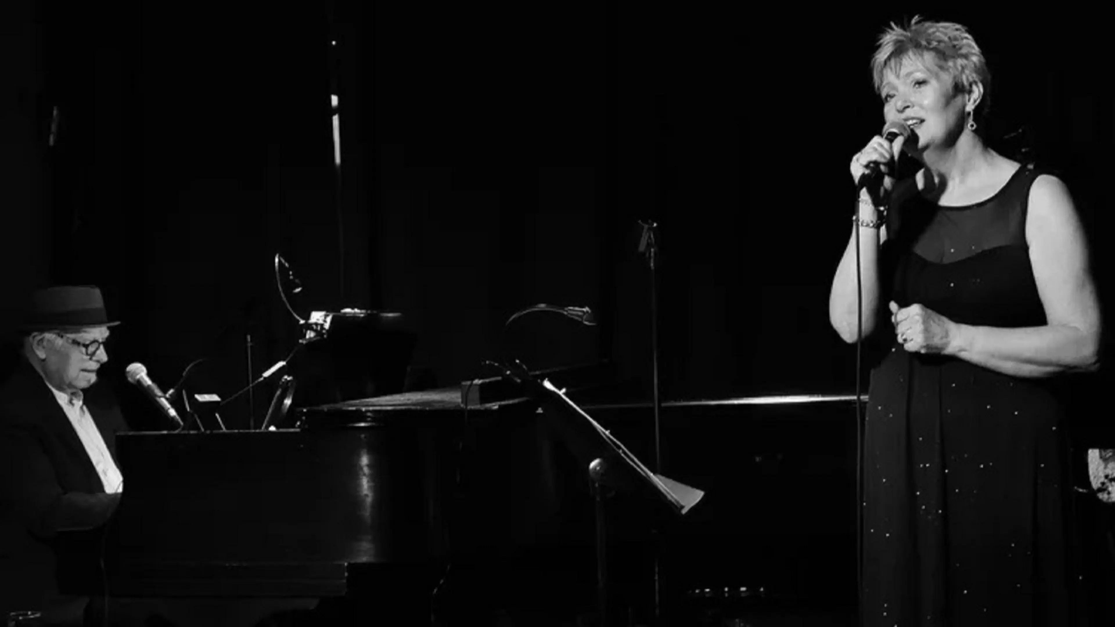 A person wearing a hat playing piano and another person in a sleeveless dress singing into a microphone on stage.