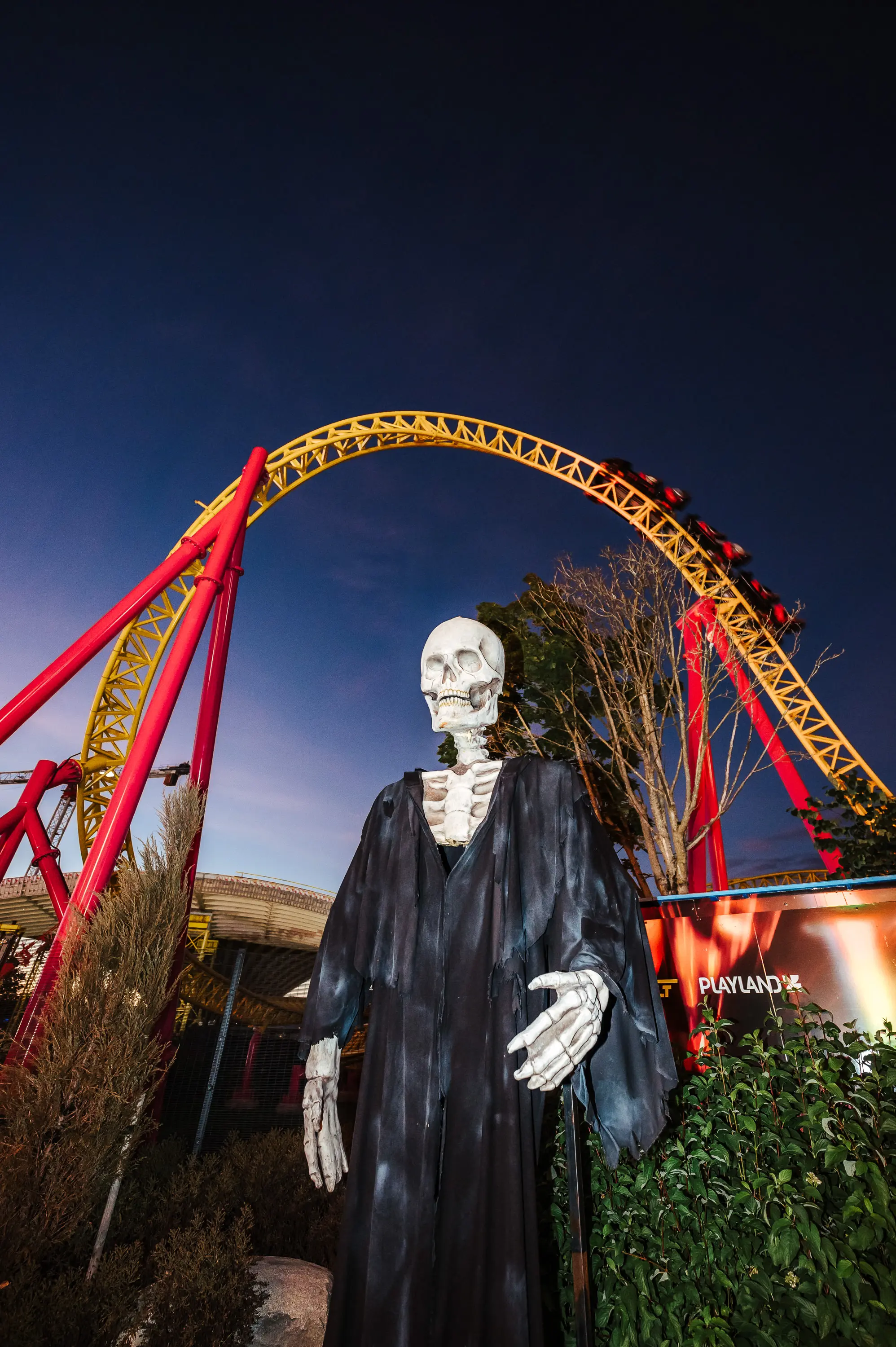Skeleton figure in a black robe standing in front of a yellow and red roller coaster at dusk.