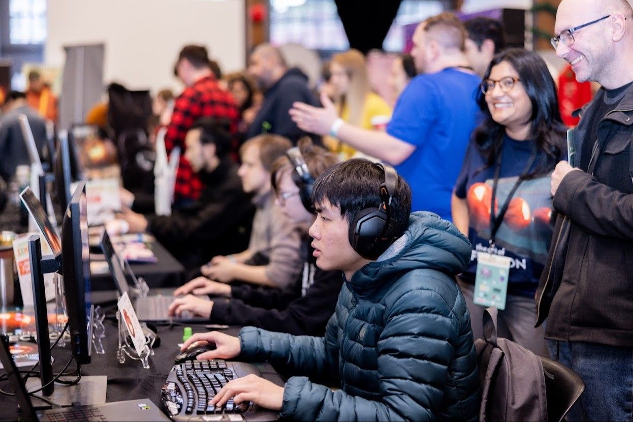 A person wearing headphones and a puffer jacket uses a keyboard and mouse at a computer station with others in the background.