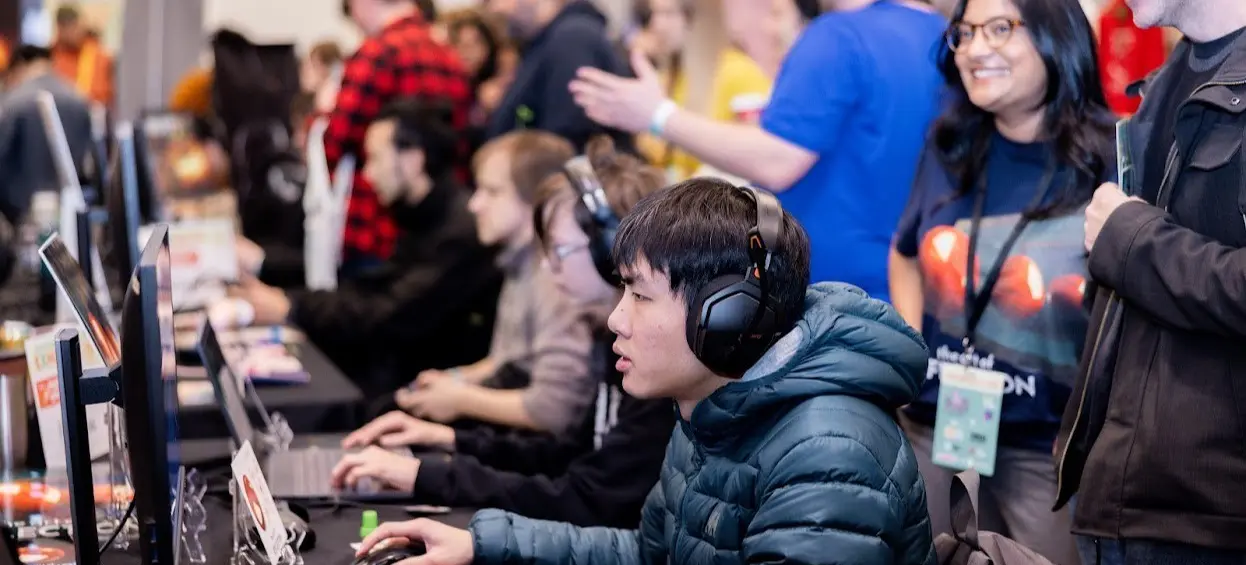 A person wearing headphones and a puffer jacket uses a keyboard and mouse at a computer station with others in the background.