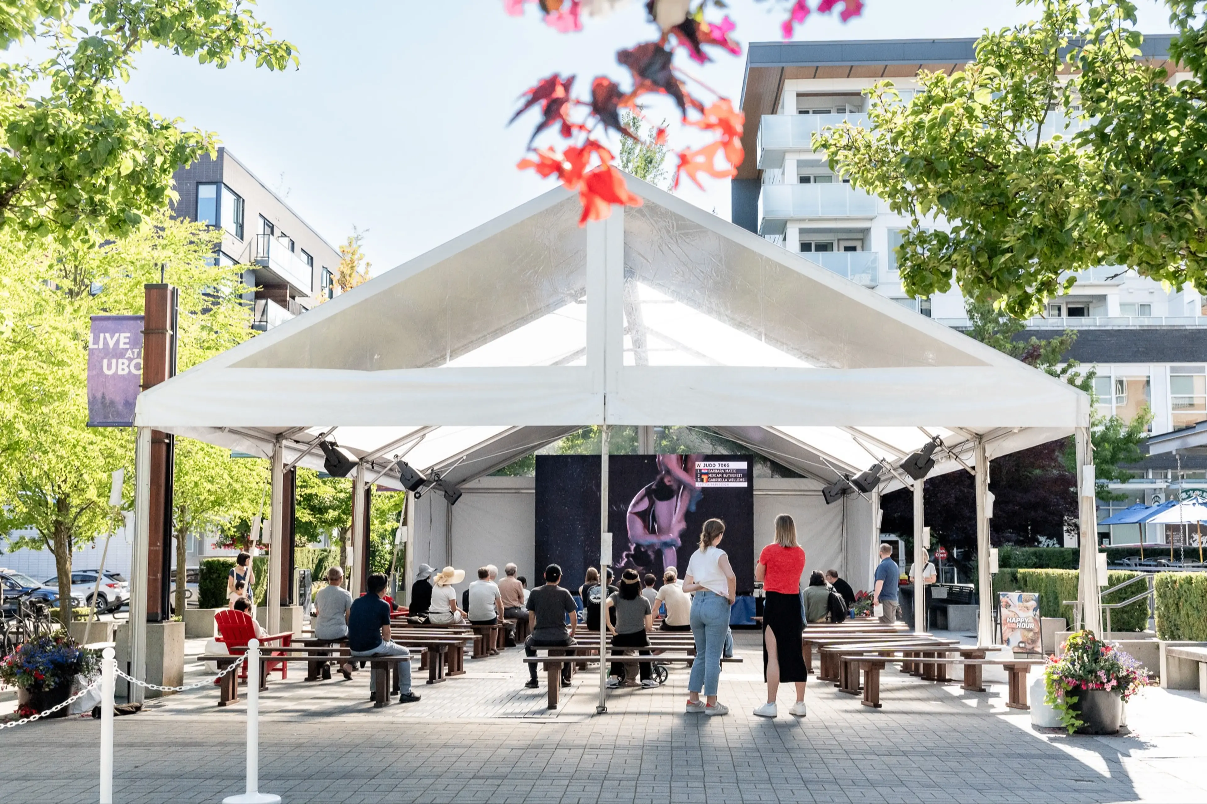 People sitting and standing under a white tent watching a large screen outdoors in a sunny urban area with trees and buildings.