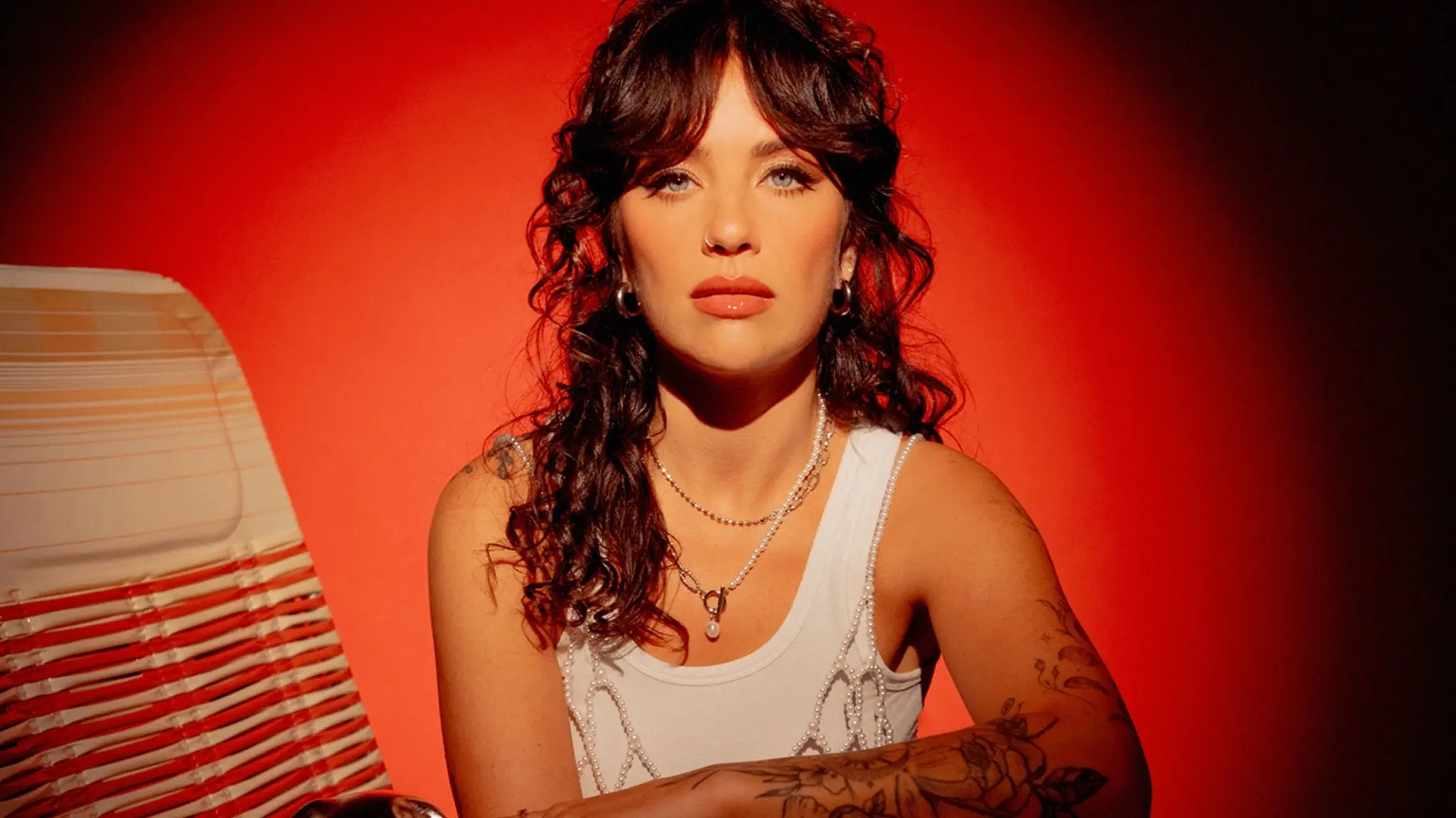 Person with curly dark hair and tattoos on arms wearing a white tank top and silver necklaces against a red background.